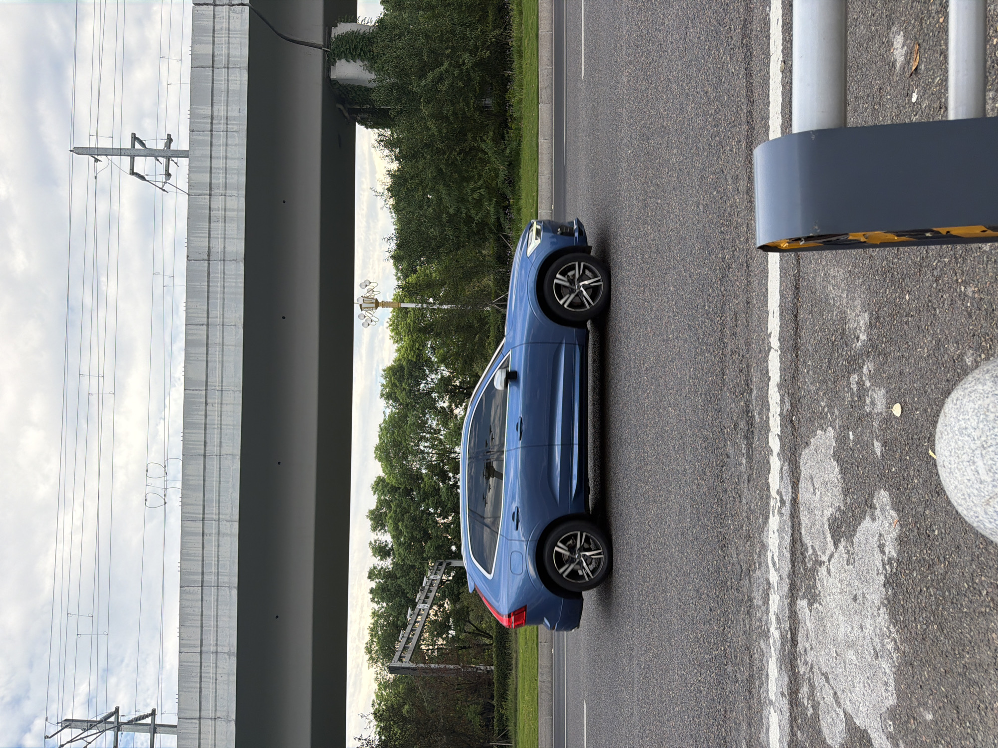 A blue Volvo XC60 is captured in profile moving along a paved road in Jiangjin, China. The car is the primary subject, positioned centrally in the frame, with its wheels on the asphalt. Overhead, a concrete overpass dominates the upper portion of the image, with power lines and utility poles visible above it. The background consists of a dense line of green trees and foliage, suggesting a suburban or semi-urban environment. The sky is overcast with scattered clouds. In the foreground, a grey and yellow traffic barrier is partially visible on the right side, and a textured concrete bollard is in the lower left corner. The road surface exhibits a white line marking the lane. The overall scene depicts urban infrastructure and vehicular traffic under a cloudy sky.