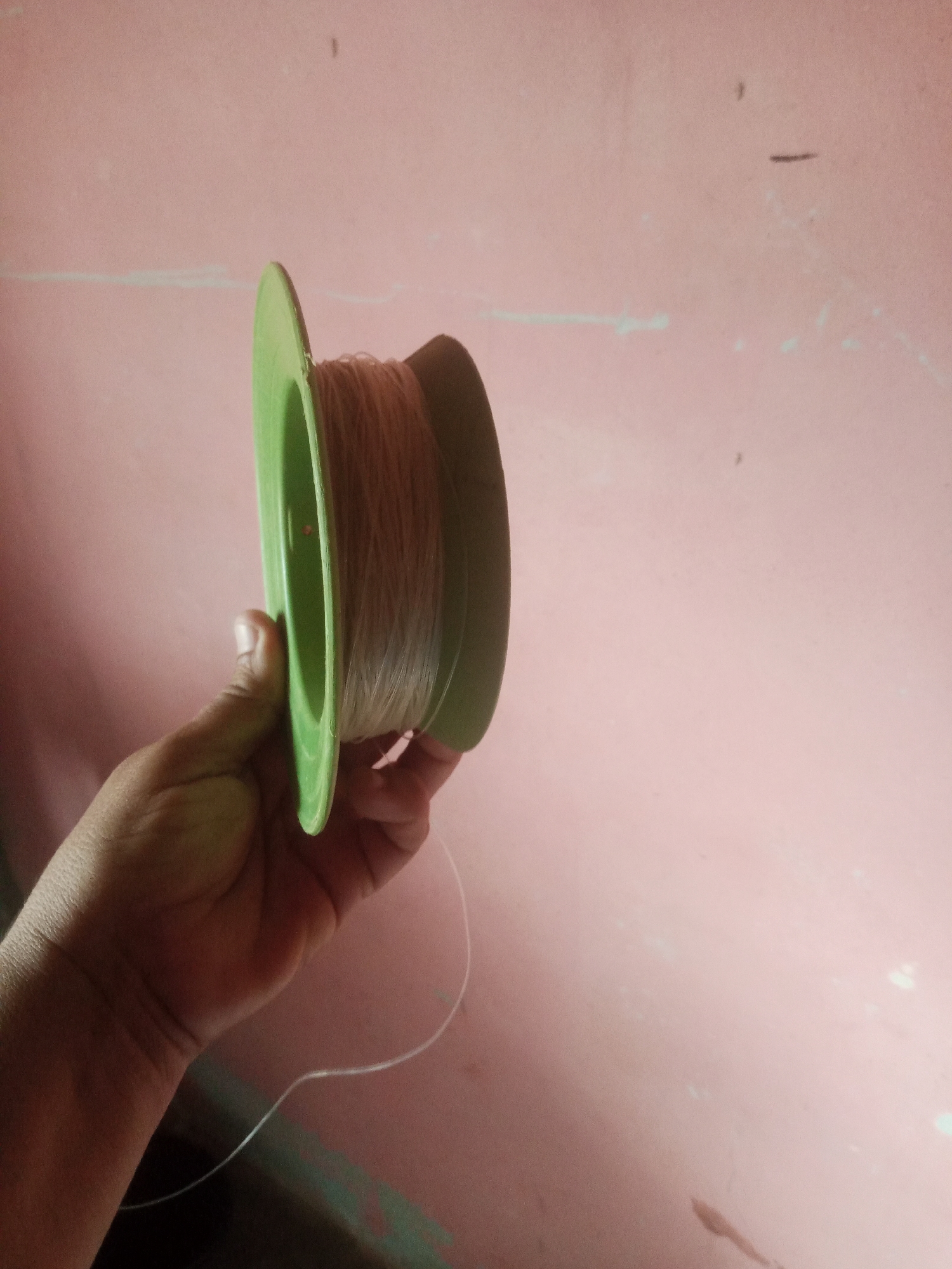 A person's hand is holding a green spool of light brown thread against a pink wall. The thread is partially unwound and trailing downwards.