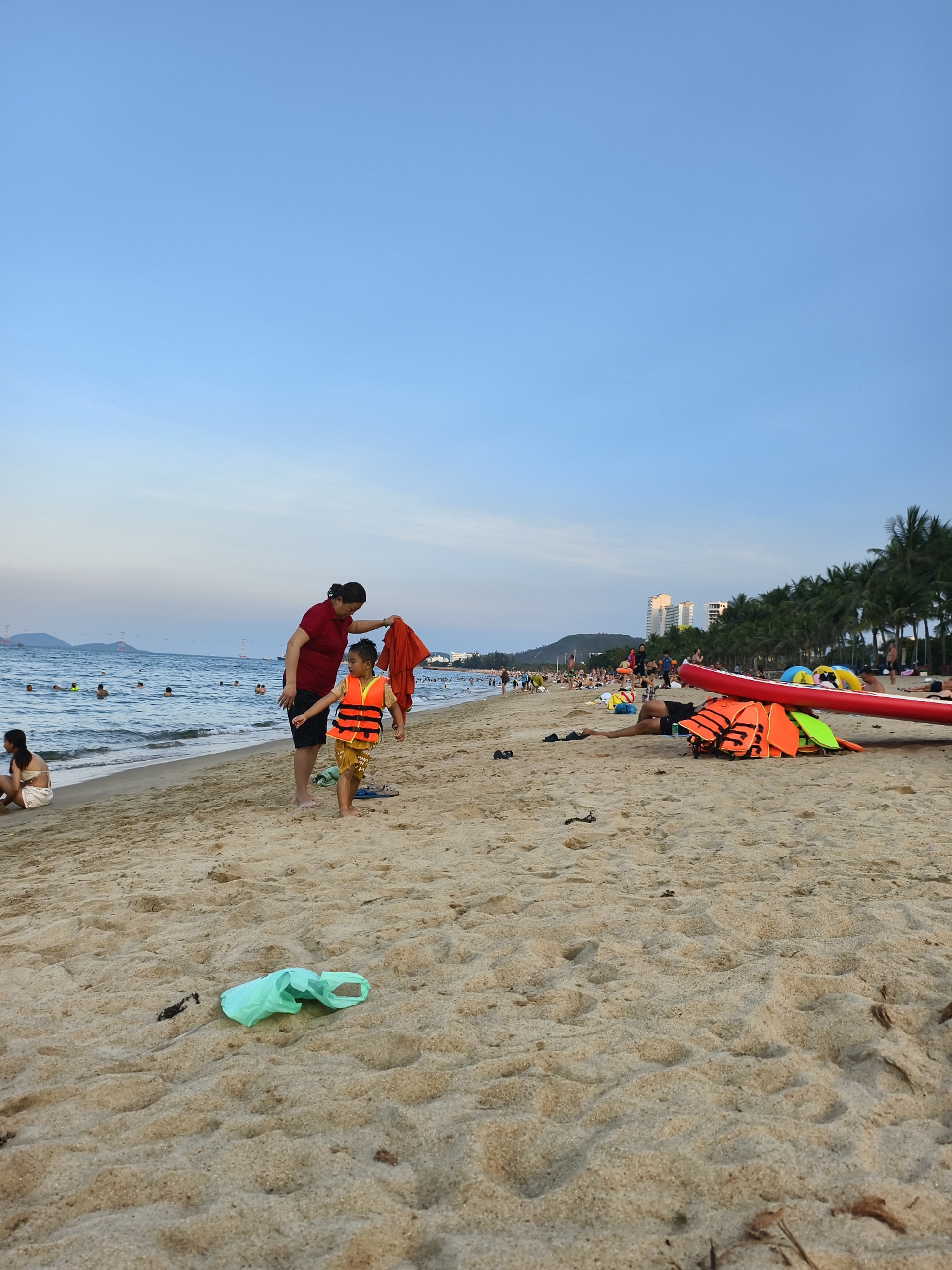 The image shows a beach scene. A woman in a red top and black shorts is drying a child who is wearing a life vest. In the background, there are people swimming and relaxing on the beach. There are also palm trees and buildings in the distance. In the foreground, there is a green plastic bag on the sand, as well as a pile of life vests and a red raft. The scene appears to be in the city of Nha Trang.