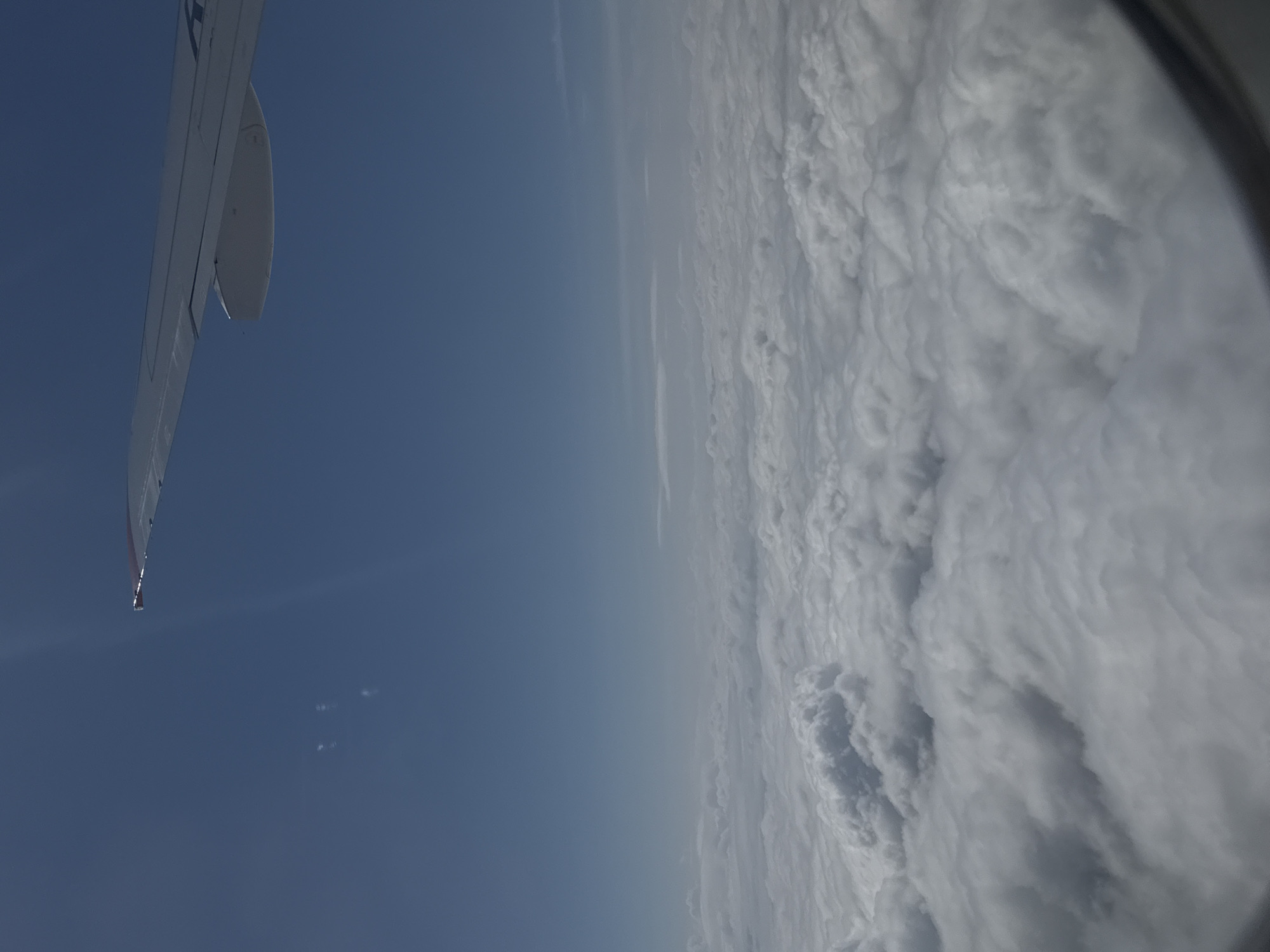 An aerial view from an airplane window shows a vast expanse of white clouds stretching to the horizon under a clear blue sky. The wing of the aircraft is visible in the upper right corner, with its tip extending into the frame. The cloud layer appears dense and textured, with variations in light and shadow suggesting depth and volume. The scene is devoid of any terrestrial features or human activity, focusing solely on the atmospheric landscape. There is no information available to connect this image to Staroe Mart'yanovo, Russia, as the image depicts a scene from an airplane at high altitude.