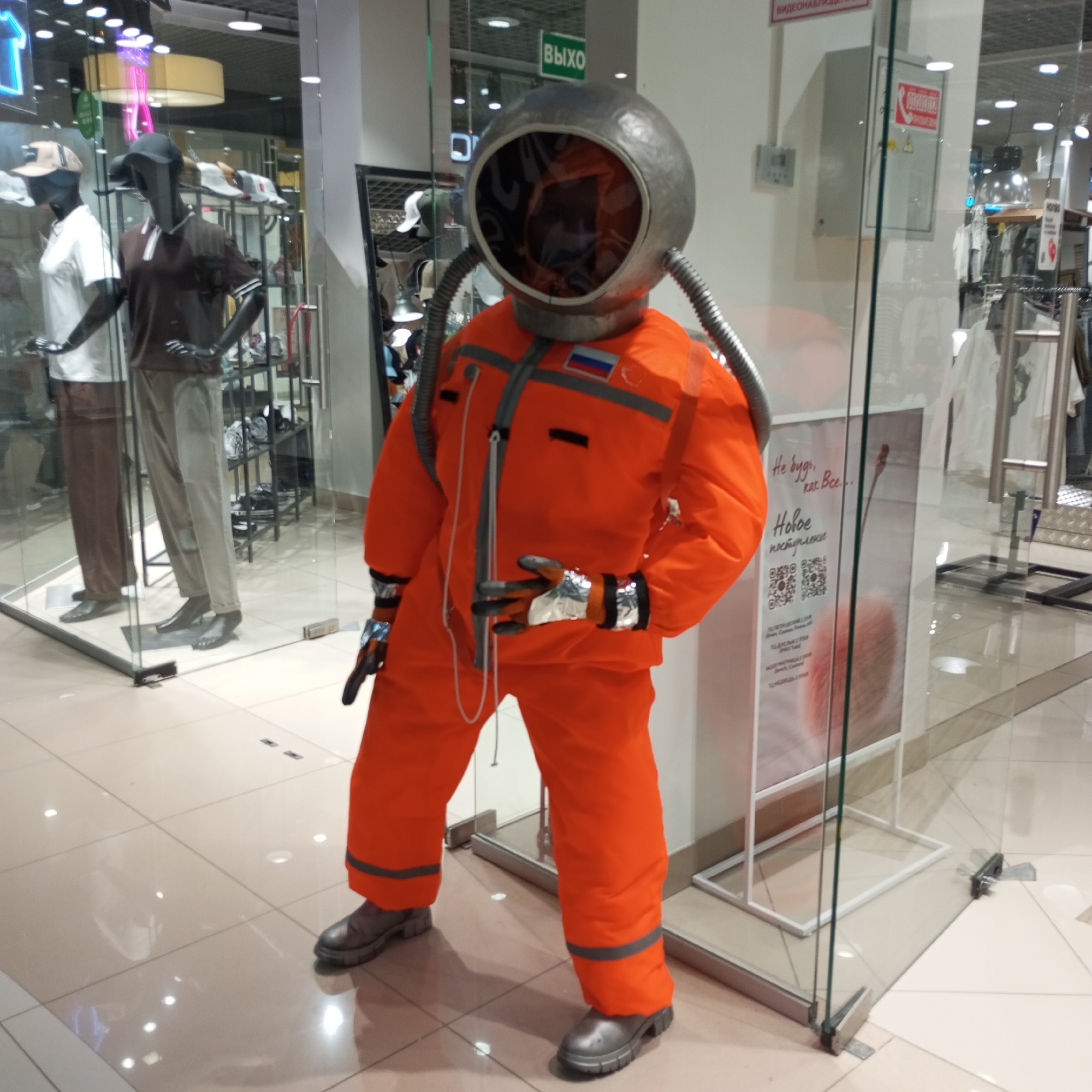 In Izhevsk, Russia, an orange astronaut suit is displayed in a storefront window. The suit, complete with a silver helmet, hoses, and reflective gloves, stands on a tiled floor in what appears to be a retail environment. The background shows mannequins dressed in casual attire and shelves stocked with merchandise, indicating it's a clothing store or department store. Glass panels form the storefront, reflecting the interior lights. A sign with Russian text is visible on the glass. The overall scene suggests a display aimed at attracting customers, possibly promoting a science fiction theme or unique apparel.