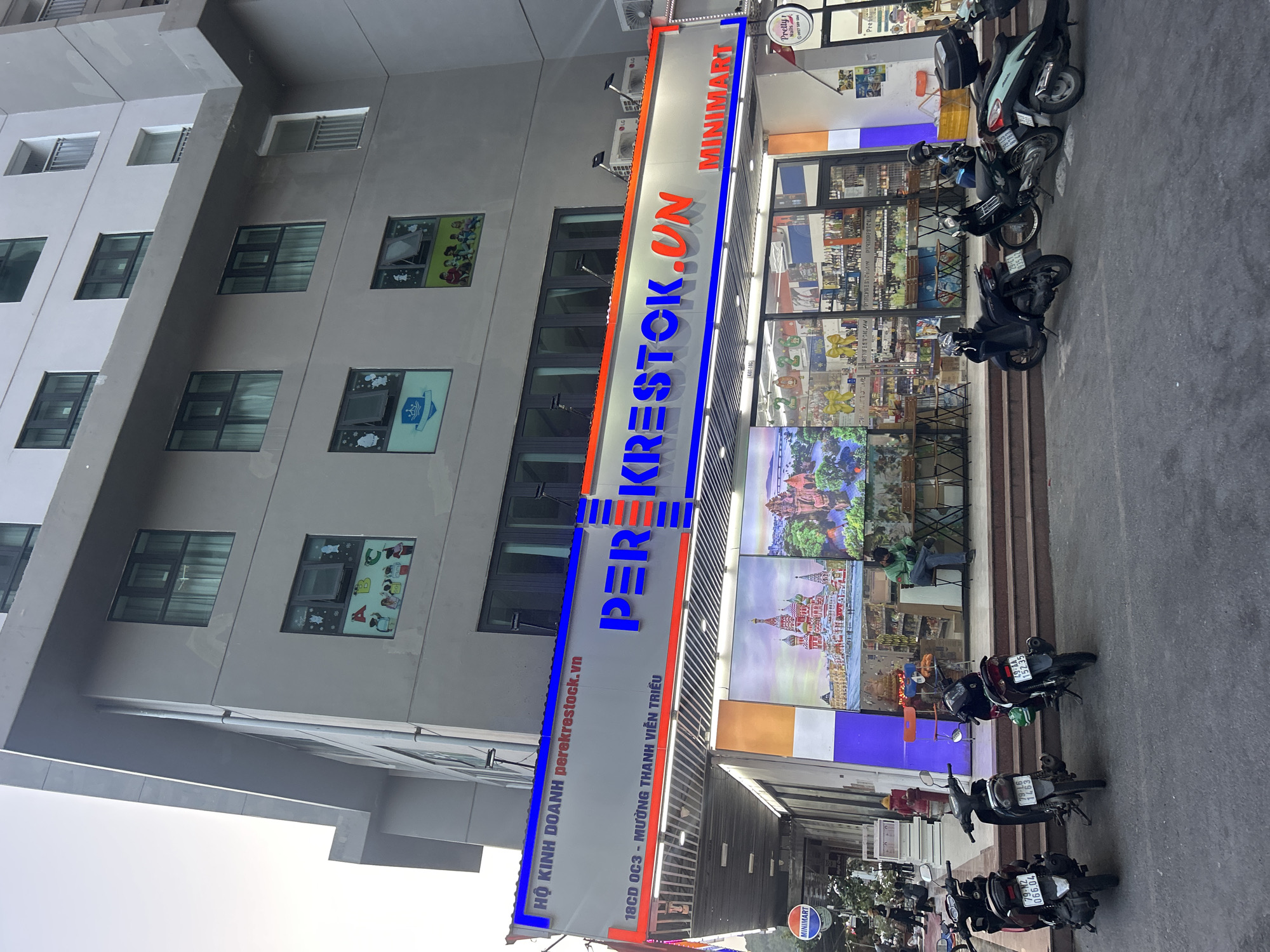 The image shows the facade of a commercial building in Bắc Nha Trang, Vietnam. The building has multiple floors and a grey exterior. Windows are visible on the left side. The most prominent feature is a large vertical sign reading "PERE KRESTOK.VN" in blue and red lettering, also featuring "MINIMART." The address, "18CD OC3 - MƯỜNG THANH VIỄN TRIỀU," is printed along the sign. The storefront displays images and products, and a person is seated inside, facing the storefront. Several motorcycles are parked on the sidewalk in front of the building, partially obscuring the view.