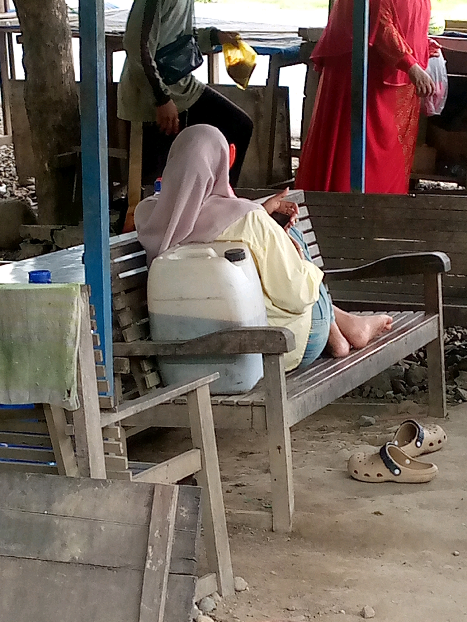 In Keudeteupinpunti, Indonesia, a scene unfolds with a focus on individuals and their surroundings. In the foreground, a wooden bench is occupied by a person wearing a pale pink hijab and a light yellow shirt, along with blue jeans. This individual is holding a mobile phone. To their left, a large white plastic container with a blue cap sits on the bench, holding a liquid with a blue tint. On the ground near the bench, a pair of beige clogs are placed. Behind this individual, another person is visible, wearing a red traditional garment. To the left, part of a table is seen, covered with a green cloth and a blue cap on top. The environment appears to be outdoors, with a rough, earthy ground surface, stones, and a wooden structure forming the background.