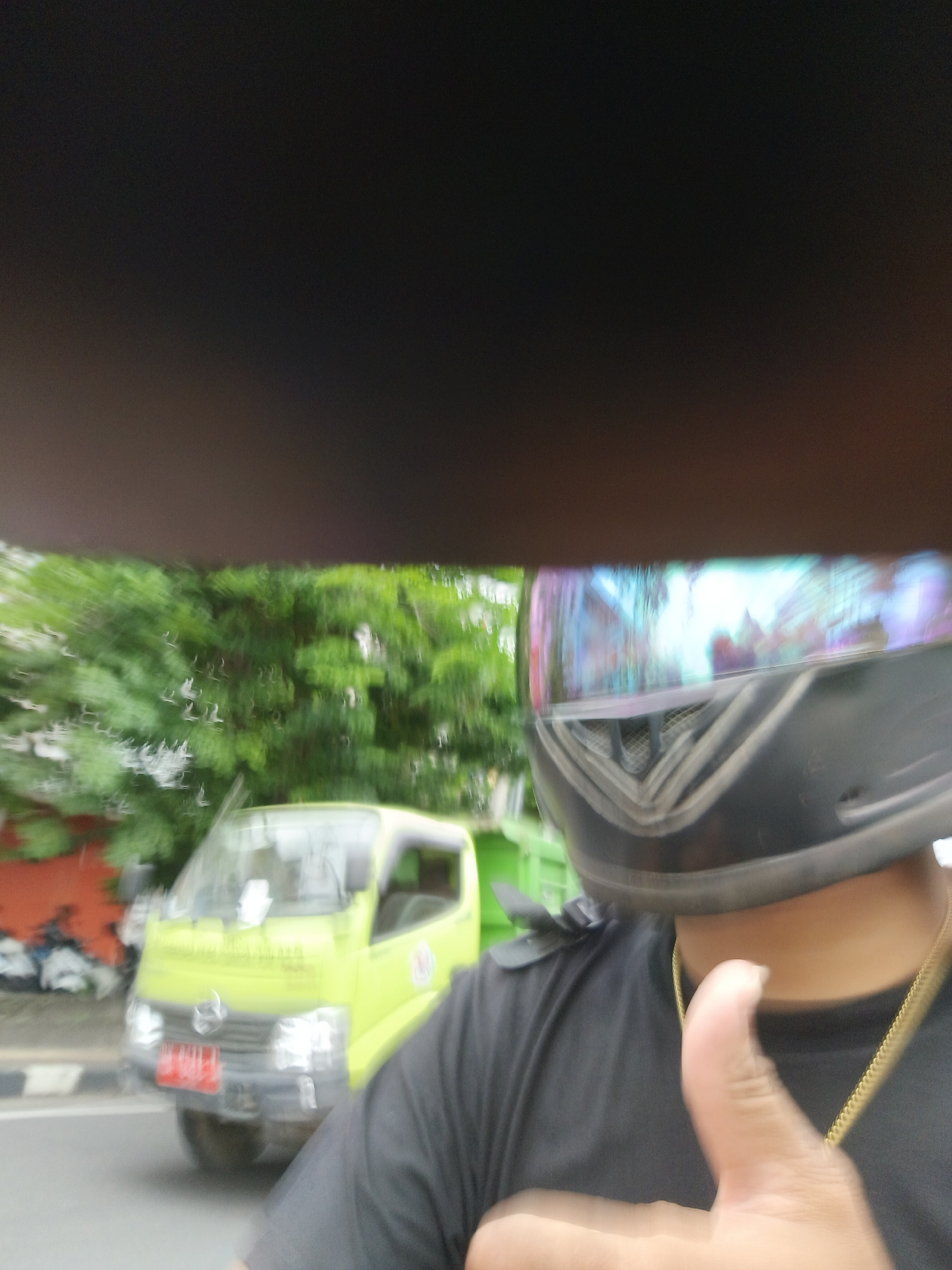 A blurry, first-person perspective shot captures a person wearing a black t-shirt and a black helmet with a reflective visor. The visor displays a colorful, iridescent reflection of the surroundings, including trees and buildings. In the background, a bright yellow truck is visible on a street. The truck has a red license plate with white characters, but they are unreadable due to the blur. The scene appears to be an outdoor environment, possibly a street in Denpasar, Indonesia, given the context. The overall image is out of focus, suggesting motion or a quick capture.