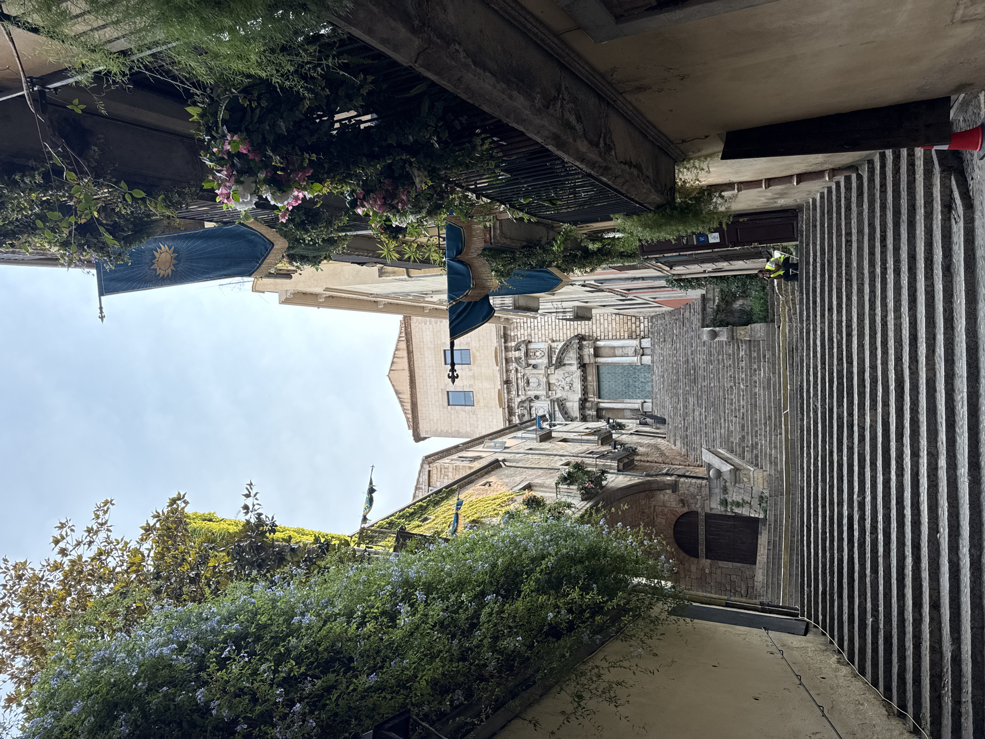 This image captures a view looking up a wide, stone staircase in the city of Girona, Spain. The staircase is flanked by buildings covered in lush green ivy and flowering vines, with blue banners featuring a sun emblem hanging from balconies. At the top of the stairs, a large building with a classical facade and a prominent green door is visible. A person wearing a yellow vest is seated on the stairs partway up, appearing to take a break. The sky is partly cloudy, and the sunlight casts shadows on the stone steps, indicating it is daytime. The overall impression is a charming, historic urban scene with an abundance of greenery.