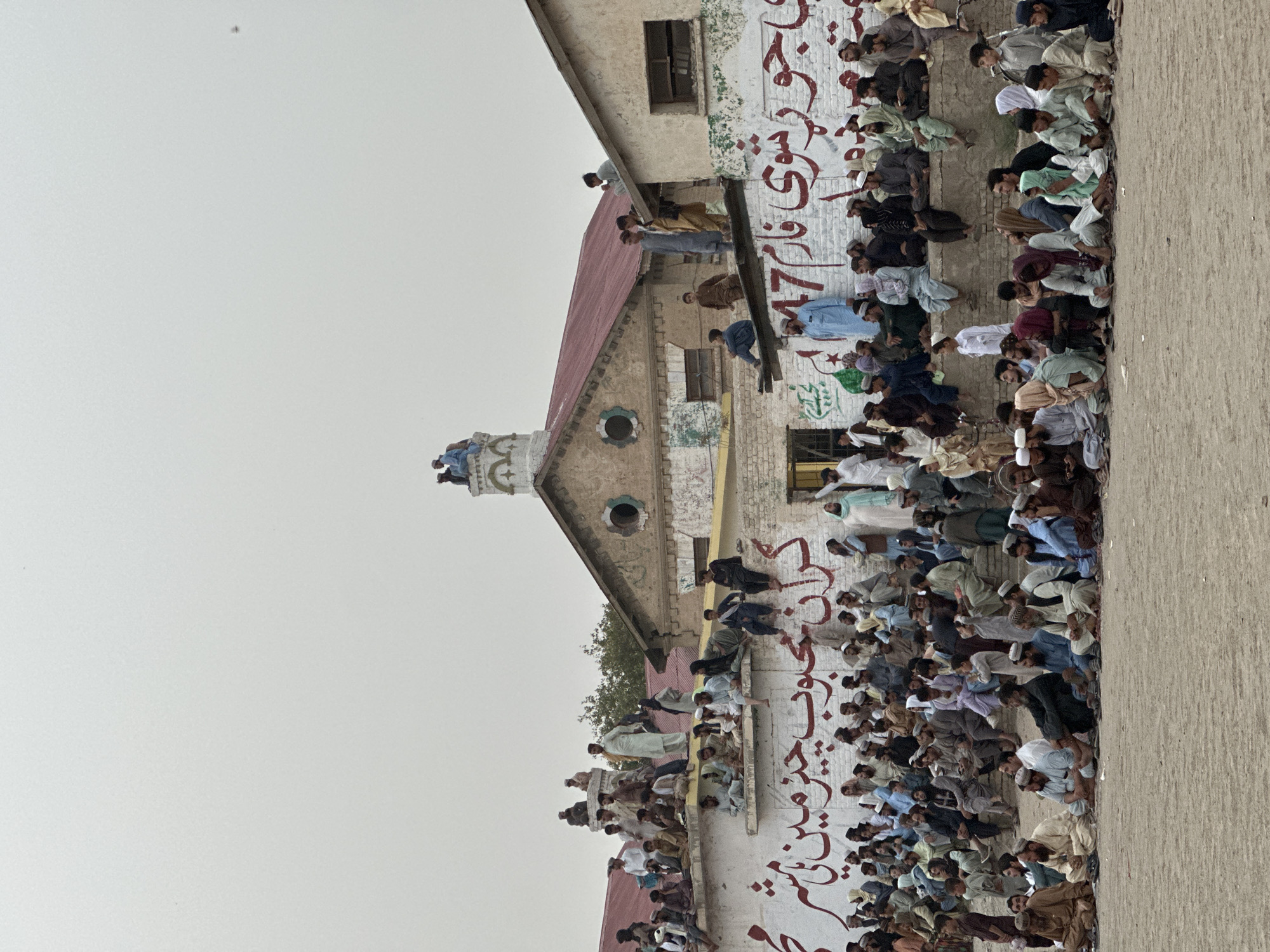 In the city of Chaman, Tehsil, Pakistan, a large gathering of men and boys is observed in front of a weathered building. The crowd is seated on the ground and on a raised concrete structure. Many individuals are wearing traditional Pakistani attire, including shalwar kameez. Some are observing a structure in the background, which appears to be a building with a red-tiled roof and a prominent clock tower-like feature. The building's facade is adorned with large red Urdu script lettering. People are also perched on the roof of the building, overlooking the scene. The overall atmosphere suggests a public event or gathering.