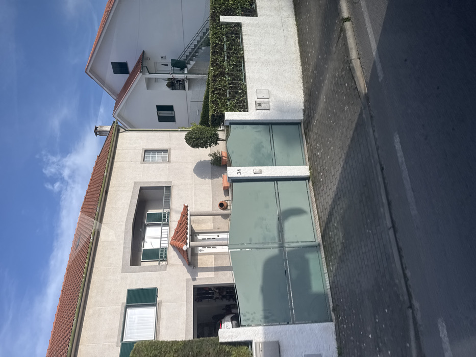 An exterior daytime photograph captures a street scene featuring two residential houses in Macedo de Cavaleiros, Portugal. The prominent house on the left is a two-story structure with a light beige stucco façade and a red terracotta-tiled roof, from which a brick chimney emerges on the right side. The ground floor includes an open garage on the far left, revealing the front portion of a white vehicle, and a central main entrance with white double doors sheltered by a small, terracotta-tiled porch roof supported by two columns. The second floor features a large window with open dark green louvered shutters on the left, a recessed central balcony with white French doors and closed dark green shutters, and a smaller window with white frames on the right. A manicured green hedge stands tall on the left, bordering the property. Near the main entrance, a small, topiary-style tree and several potted plants are visible.
To the right of the primary house, a second, white-stuccoed residence with a steeply pitched red terracotta-tiled roof is present. This house features an external metal staircase leading to an upper-level entrance and at least two windows, one of which has closed dark green shutters.
The foreground displays a large, closed, double-leaf gate in a sage green color, horizontally segmented, set between two white stucco pillars. The right pillar is marked with the number "34" and contains an intercom unit. Extending to the right from this pillar is a low white stucco wall topped with a dense, green hedge, which also features two utility boxes. The property is separated from the street by a sidewalk paved with dark grey interlocking bricks. An asphalt street with white dashed lines occupies the immediate foreground. Bright sunlight from the left casts long shadows from the gate and houses across the sidewalk and street. No individuals are present in the scene.