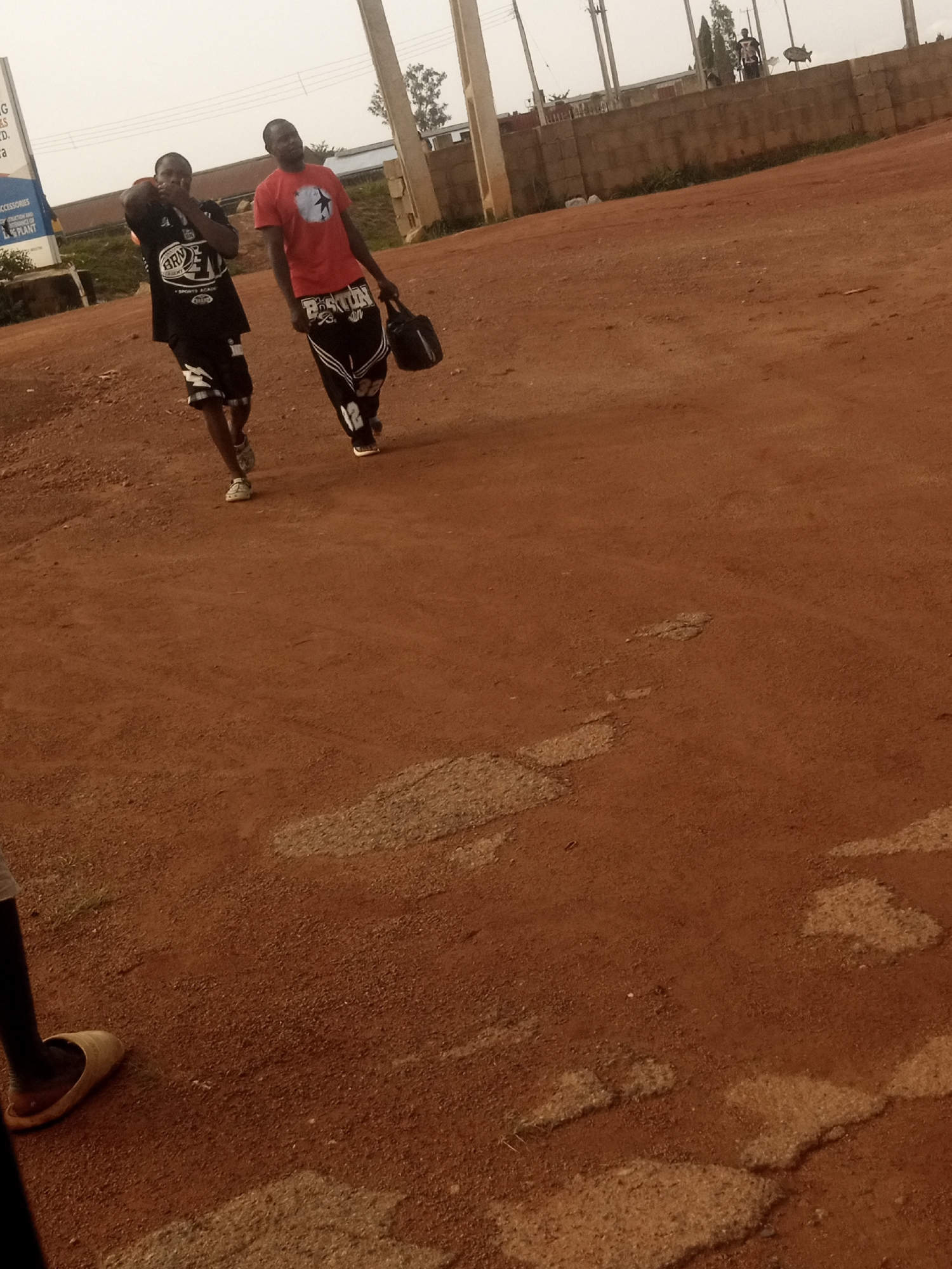 Two young men walk on a dirt road in what appears to be a developing area. The man on the left wears a black t-shirt with white lettering that appears to say "BRN ACADEMY" and black shorts. The man on the right wears a red t-shirt with a white star design and black pants with white stripes and the number "12" in white. He is carrying a black bag. The ground is red dirt with some lighter patches. In the background, there is a concrete wall, some trees, and utility poles. A sign is visible on the left side of the image, with some text in English. The sky is overcast.