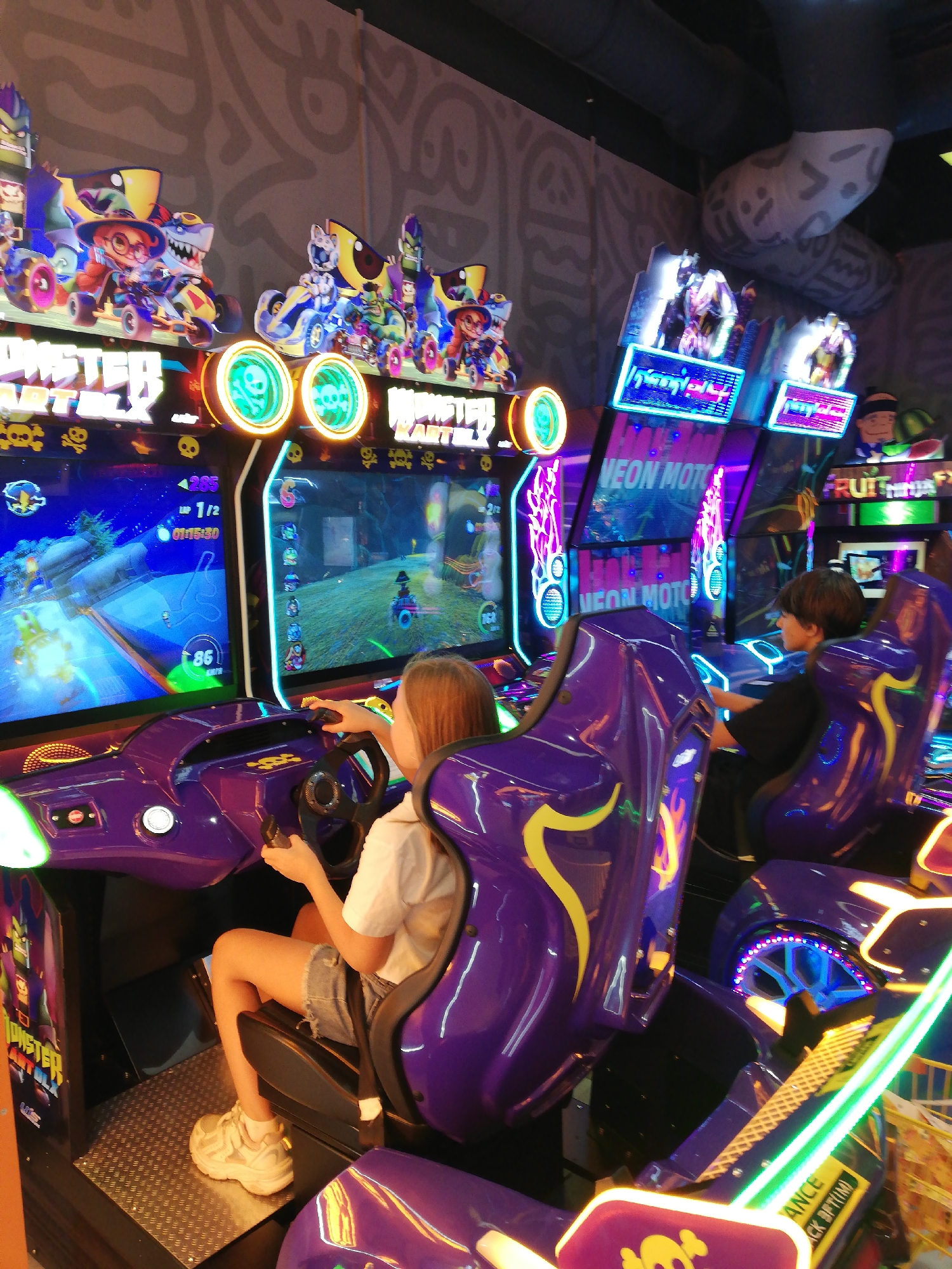 This image depicts the interior of an arcade in Saint Petersburg, Russia, likely a family entertainment center or a dedicated arcade. The focus is on a row of brightly lit, purple and yellow racing arcade game cabinets. In the foreground, a young girl with blonde hair, wearing a white t-shirt and denim shorts, is seated in one of the cabinets, her hands on the steering wheel. She is playing a racing game with cartoonish characters visible on the screen, displaying elements like speed and lap count. To her right, a boy with brown hair, wearing a black t-shirt, is seated in another adjacent cabinet, also playing a game. The arcade environment is characterized by its vibrant, neon lighting, with the games themselves featuring flashing lights and colorful graphics, including skulls and flames. The overall impression is one of a lively and engaging entertainment space.