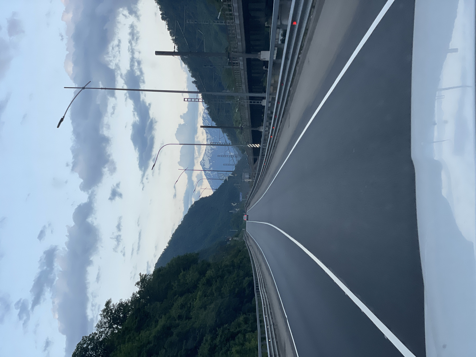 The image captures a view from a vehicle driving on a paved road in Chvizhepse, Russia. The road is divided by white lines, indicating multiple lanes. On the left side of the road, a steep, tree-covered mountainside rises. On the right, a metal guardrail runs along the edge of the road, and beyond that, an elevated structure, possibly a bridge or viaduct with train tracks, is visible. In the distance, snow-capped mountains are visible under a cloudy sky. A single white car is seen driving ahead on the road. Streetlights are positioned along the road.