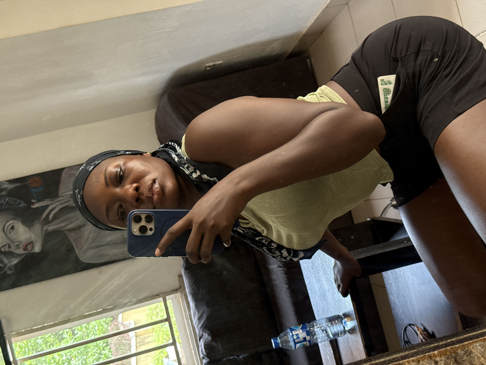 A woman is taking a selfie in what appears to be a bathroom in Abuja, Nigeria. She is wearing a headscarf, a light green t-shirt, and black shorts, with what looks like cash visible in her shorts pocket. She is holding a blue smartphone with multiple cameras. Behind her, there is a painting of a woman and a window with a view of green trees. A bottle of water is seen on a dark surface next to her.
