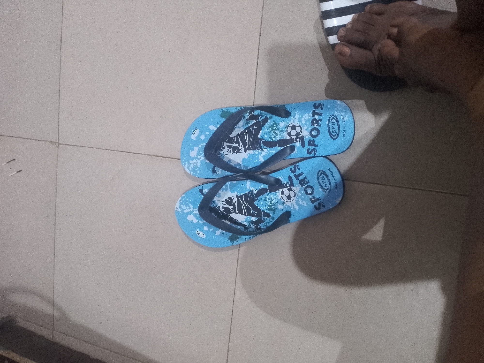 An overhead, medium shot captures a pair of blue flip-flops with black straps, each adorned with a graphic of a soccer player in mid-kick and the word "SPORTS" in bold white lettering. The flip-flops are positioned on a light-colored, tiled floor, with one slightly overlapping the other. To the upper right of the flip-flops, a person's foot is visible, wearing a black and white striped sandal. The floor shows subtle grout lines between the tiles, and a soft shadow is cast to the right of the flip-flops and foot, suggesting the presence of a light source from the left. The overall environment appears to be an indoor setting with neutral-toned flooring.