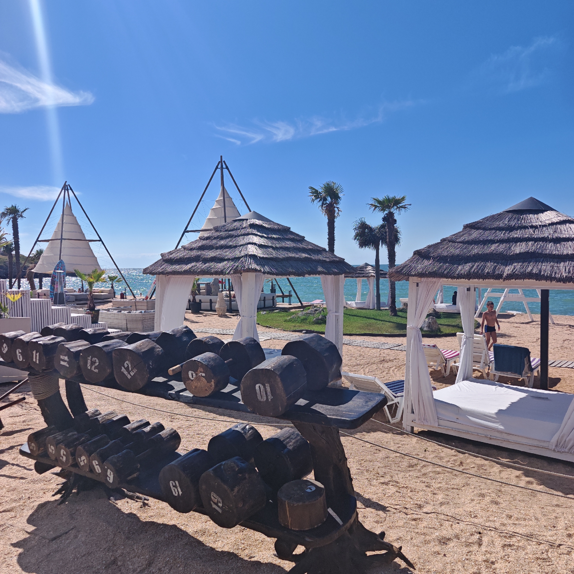 The image depicts a sunny day at a beachside resort in Мысовское сельское поселение, Ukraine. In the foreground, a wooden rack holds a collection of large, rustic dumbbells made of logs, some marked with numbers like '6', '11', '12', and '10'. The beach is covered in fine sand. Behind the dumbbells, thatched-roof cabanas with white curtains offer shade, with some containing white lounge beds. In the middle ground, the turquoise ocean stretches to the horizon under a clear blue sky with a few wispy clouds. Palm trees dot the landscape, and in the distance, more resort structures and lounge areas are visible. A young boy in blue swim trunks walks on the sand near one of the cabanas. The overall scene conveys a relaxed vacation atmosphere with a unique, outdoor fitness element.
