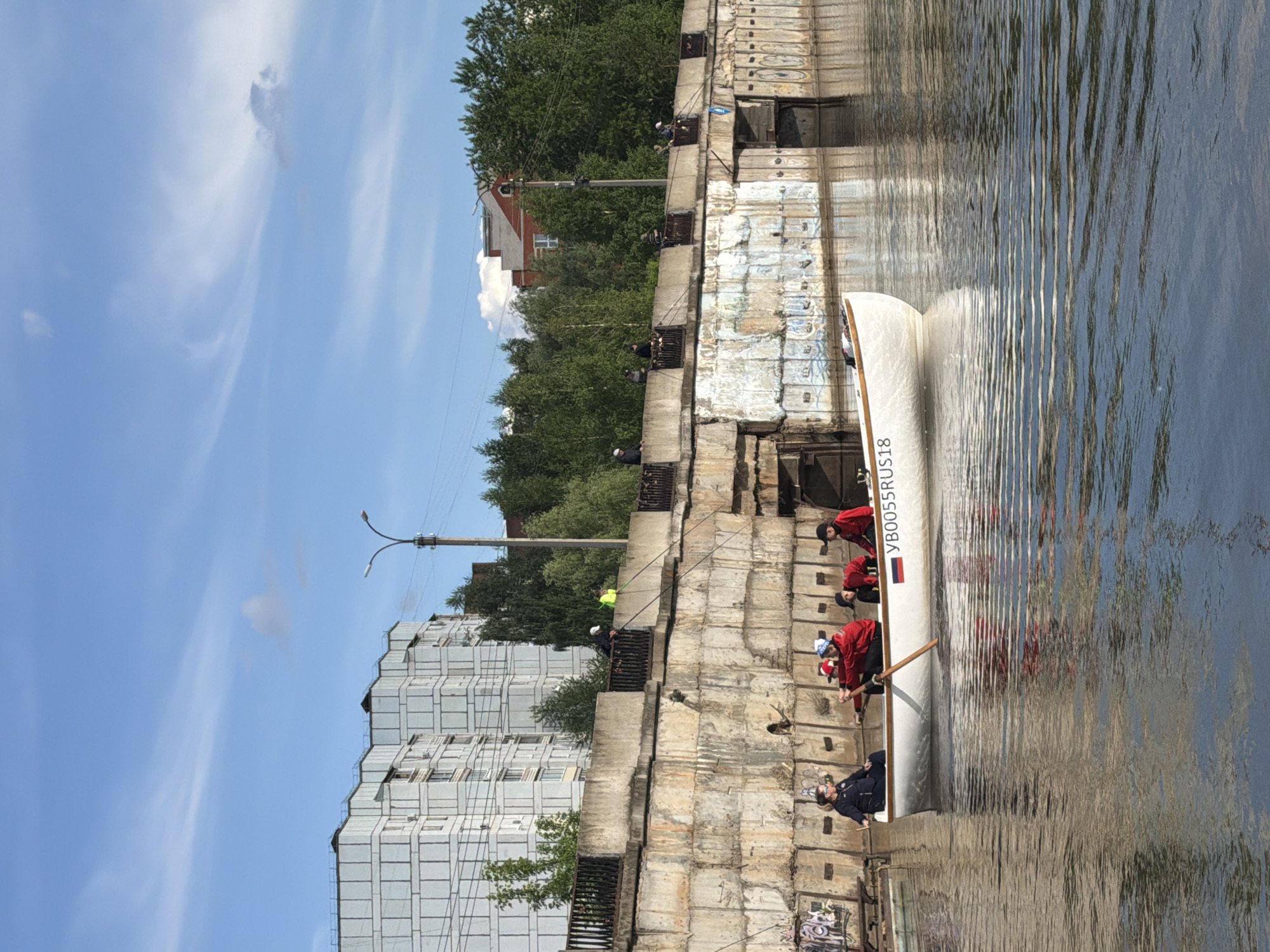 A group of people are rowing on a river in Votkinsk, Russia. The river runs along a concrete embankment that supports building structures above.
The boat is a white, long canoe with the inscription "YB0055RUS18" and a small Russian flag on its side. A team of four people are rowing it, all dressed in red jackets and hats, plus a supervisor in the front, dressed in what appears to be a security uniform of black with blue stripes.
The embankment above is made of stacked concrete blocks and grates, with a group of fishermen along its edge, fishing into the river.
On the opposite side of the embankment are trees, a building, and a streetlight. The sky is a bright blue with wispy clouds.