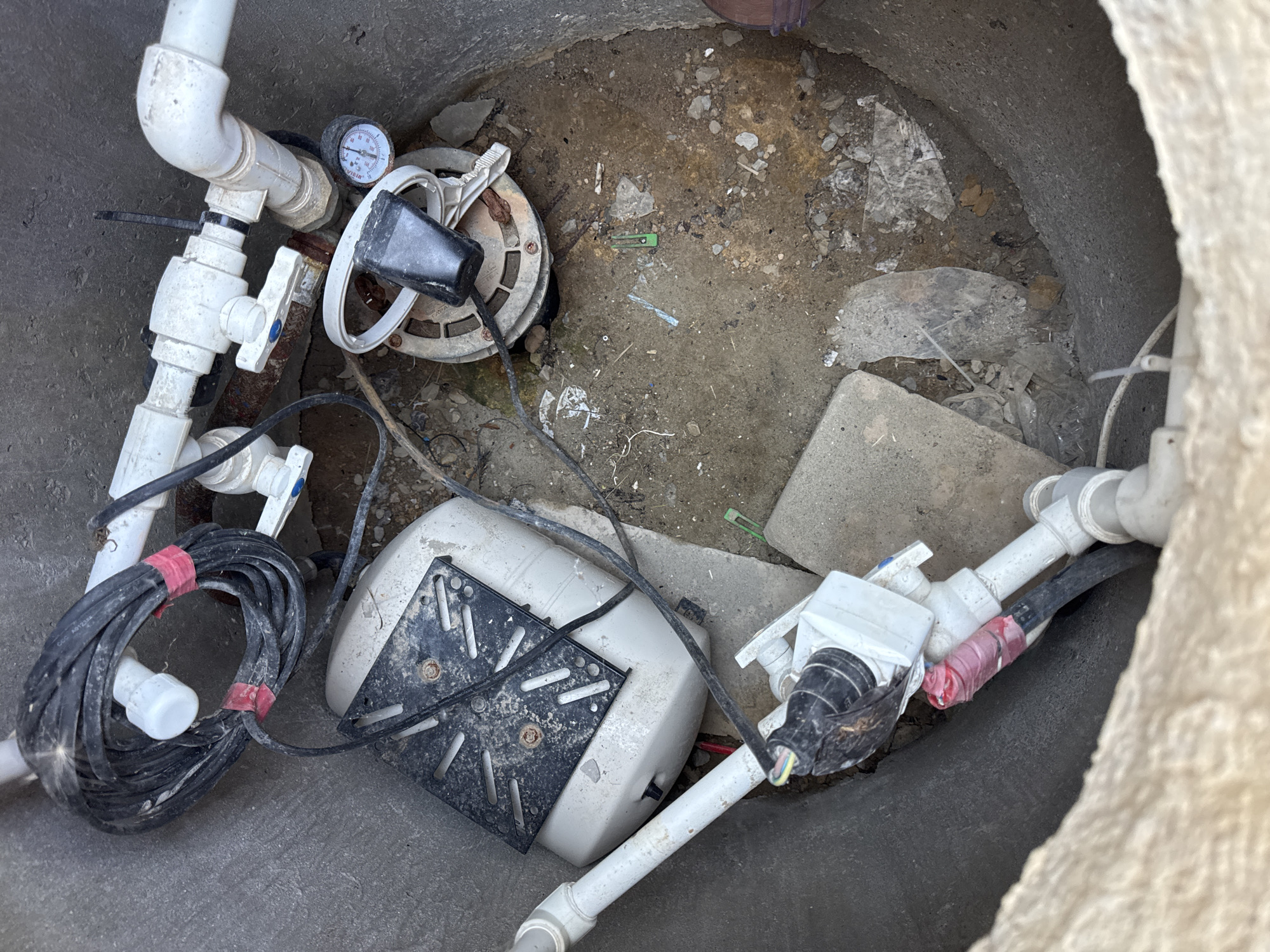 The image shows a round, concrete pit in Uyutnoye, Ukraine, containing plumbing equipment. In the center of the pit is a white pressure tank with a black metal bracket on top. To the right of the tank is a circular component with a pressure gauge attached to a white pipe. Multiple white PVC pipes and a black electrical cable are visible throughout the pit, some secured with red tape. The pit floor is covered in dirt, small rocks, and debris.
