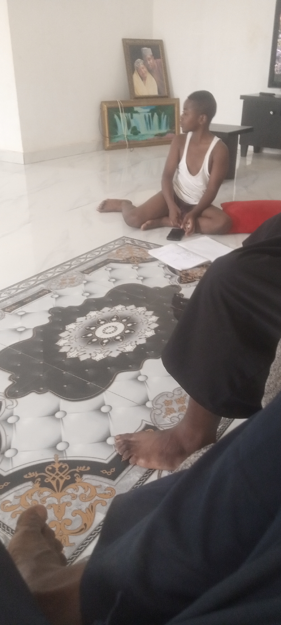 A photo taken indoors in Narayi, Nigeria. A young, dark-skinned person wearing a white tank top sits cross-legged on a tile floor, with a red pillow behind them and a black mobile phone and an open book in front. Behind the young person is a white wall, with a picture of an older couple and a painting of a waterfall hanging on it, along with a black table and TV stand and a television. In the foreground are the bare feet and legs of a person wearing dark pants. The floor is covered in large tiles with a decorative pattern.