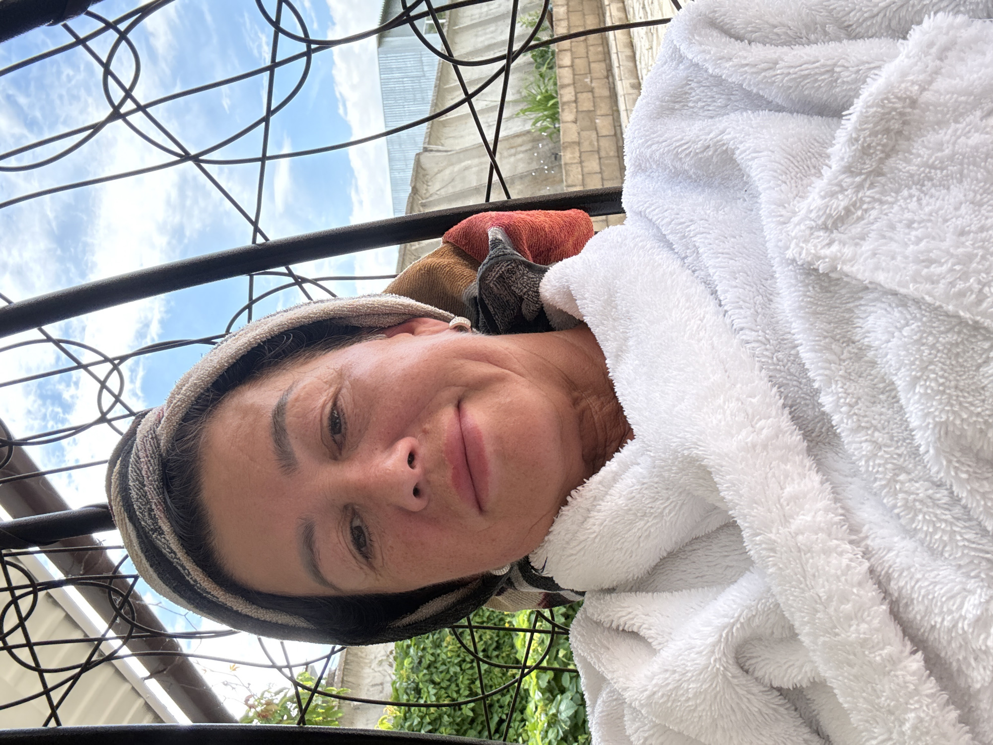 A selfie of a woman taken outdoors. She is wrapped in a white terrycloth robe and has a striped headband on her head. The woman is smiling at the camera and appears to be relaxed. Behind her, a black metal railing is visible, and beyond that, a blue sky with white clouds, some greenery, and a brick building can be seen. The image is oriented vertically. The lighting suggests it is daytime. The presence of the robe and headband might indicate the woman is preparing to go out, has just returned, or is simply enjoying a leisurely moment outdoors. The background details suggest a residential or garden setting.