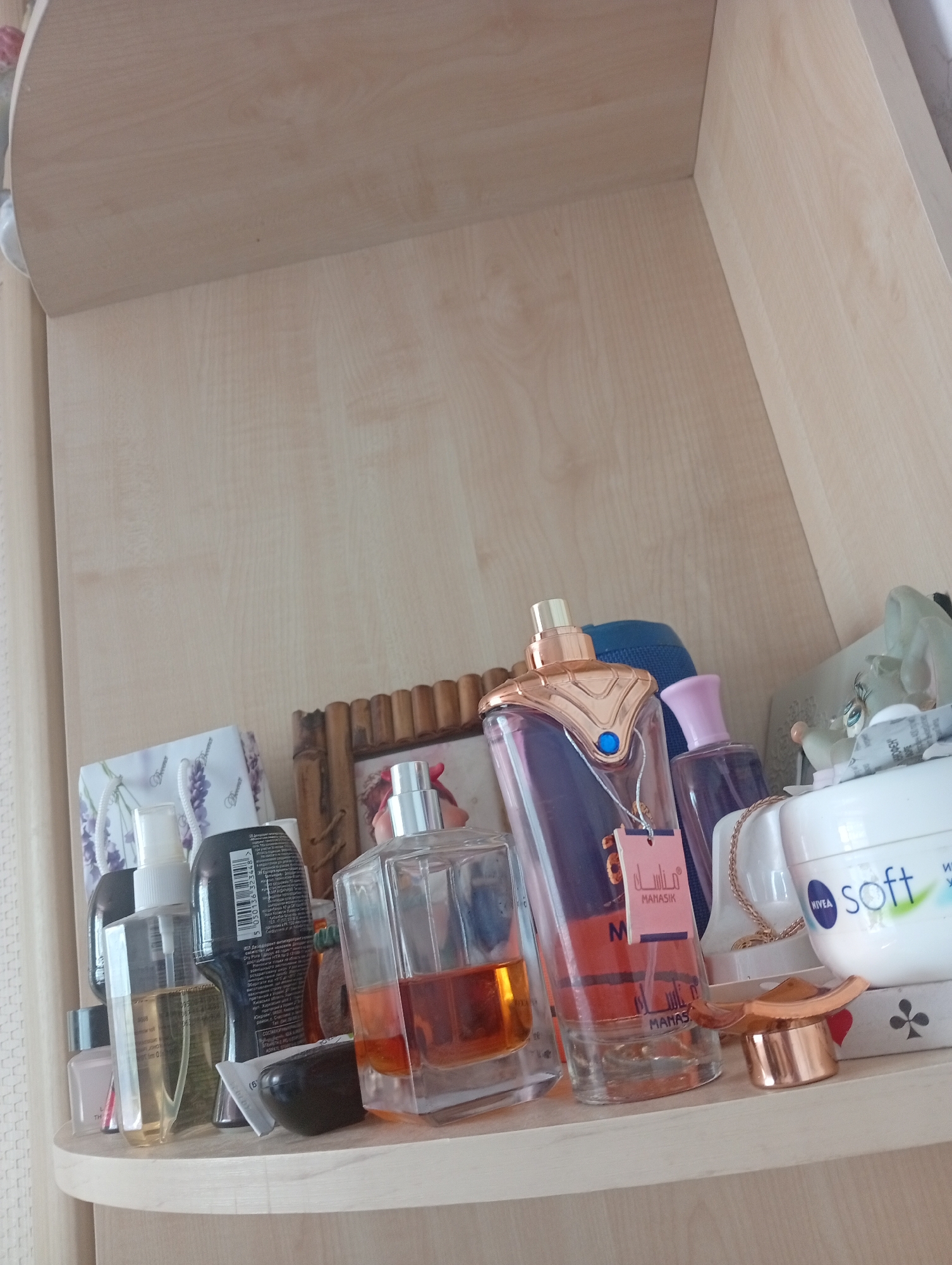 The image shows a shelf in what appears to be a domestic setting, likely a bathroom or dressing area, in Mykolaiv, Ukraine. The shelf is made of light-colored wood. Several cosmetic and perfume bottles are arranged on the shelf.
On the left side, there are smaller bottles including a clear bottle with a white pump dispenser containing a light yellow liquid. Next to it is a dark, cylindrical bottle with a label containing text in Cyrillic script. A pink nail polish bottle is partially visible at the far left.
In the center of the shelf, a prominent perfume bottle with a unique gold-colored cap and a dark purple glass body is visible. A small pink tag is attached to the bottle. To the right of this bottle is another perfume bottle with a pink cap and a clear glass body containing a purple liquid. Behind these bottles, a blue fabric item is partially visible.
Further to the right, a square glass perfume bottle with a light orange liquid is visible. Next to it is a white jar with blue and green text that reads "NIVEA soft". A gold-colored chain and a small gold-colored object resembling a heart or clover are placed in front of the NIVEA jar. A framed object with a wooden border is positioned behind the perfume bottles, its contents are partially obscured but appears to be a photograph or decorative item. To the right of the NIVEA jar, a light green figurine, possibly a character, is visible.
The background of the image is the wooden shelf and the wall behind it, which also appears to be made of light-colored wood paneling. The lighting suggests an indoor environment. No people or actions are depicted.