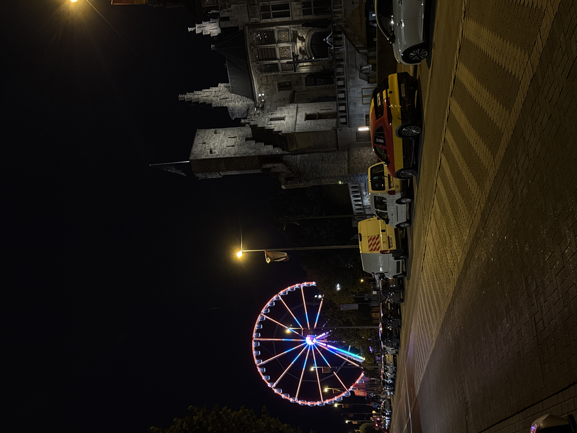 A night scene depicts a brightly lit Ferris wheel with red, white, and blue lights, situated in the background on the left side of the image. In the mid-ground, a row of parked cars lines the street, with a white van prominently displayed in the center right. Adjacent to the street is a cobblestone sidewalk. Behind the cars and Ferris wheel, a large, ornate stone building with a tall tower is visible, illuminated by lights. The sky is dark, indicating nighttime. The scene is likely in Antwerp, Belgium, given the building's architecture and the presence of a Belgian flag on the tower.