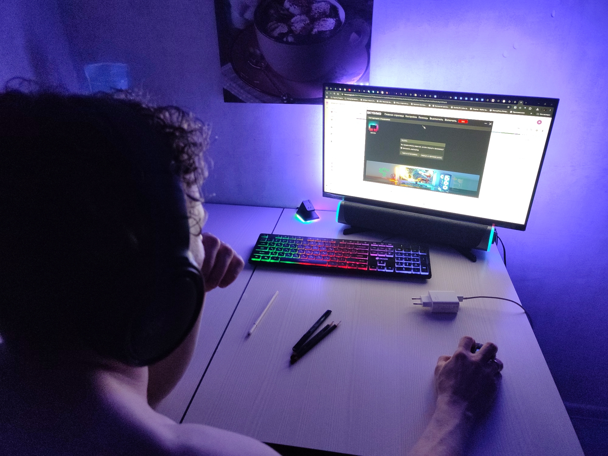 In a dimly lit room with purple and blue ambient lighting, a person wearing headphones is seated at a desk in front of a computer monitor. The person is viewed from behind and to the side, with their right hand resting on a mouse. The desk is made of a light-colored wood-grain material and has a keyboard with colorful, illuminated keys. Several writing utensils and a white object, possibly a stylus, are also on the desk. A white power adapter is plugged into an unseen outlet, with its cord running towards the computer. The computer screen displays a webpage with Russian text and images. In the background, a framed artwork hangs on the wall, and the room appears to be a personal workspace or gaming setup.