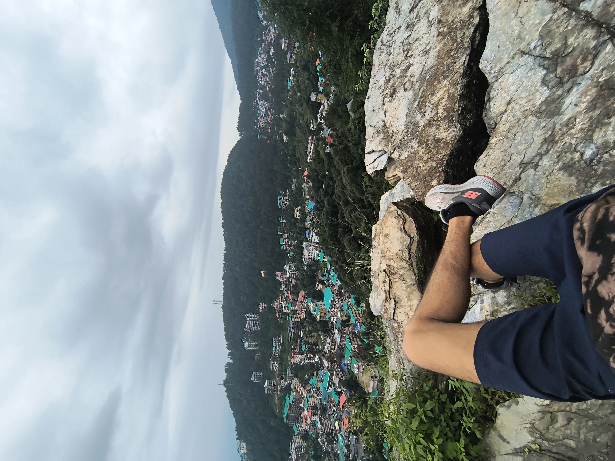 A first-person perspective shot shows a person sitting on rocks overlooking a cloudy, mountainous cityscape in Bhagsu, India. The individual's legs, clad in navy blue shorts and a patterned t-shirt, are crossed as they wear grey athletic shoes with pink accents. The foreground is dominated by the rough, textured surface of large rocks, with green foliage peeking out from the left side. In the middle ground, a densely packed town with multi-story buildings, many featuring turquoise roofs, sprawls across the lush, green hillside. The background consists of a heavily forested mountain slope under a thick, overcast sky.