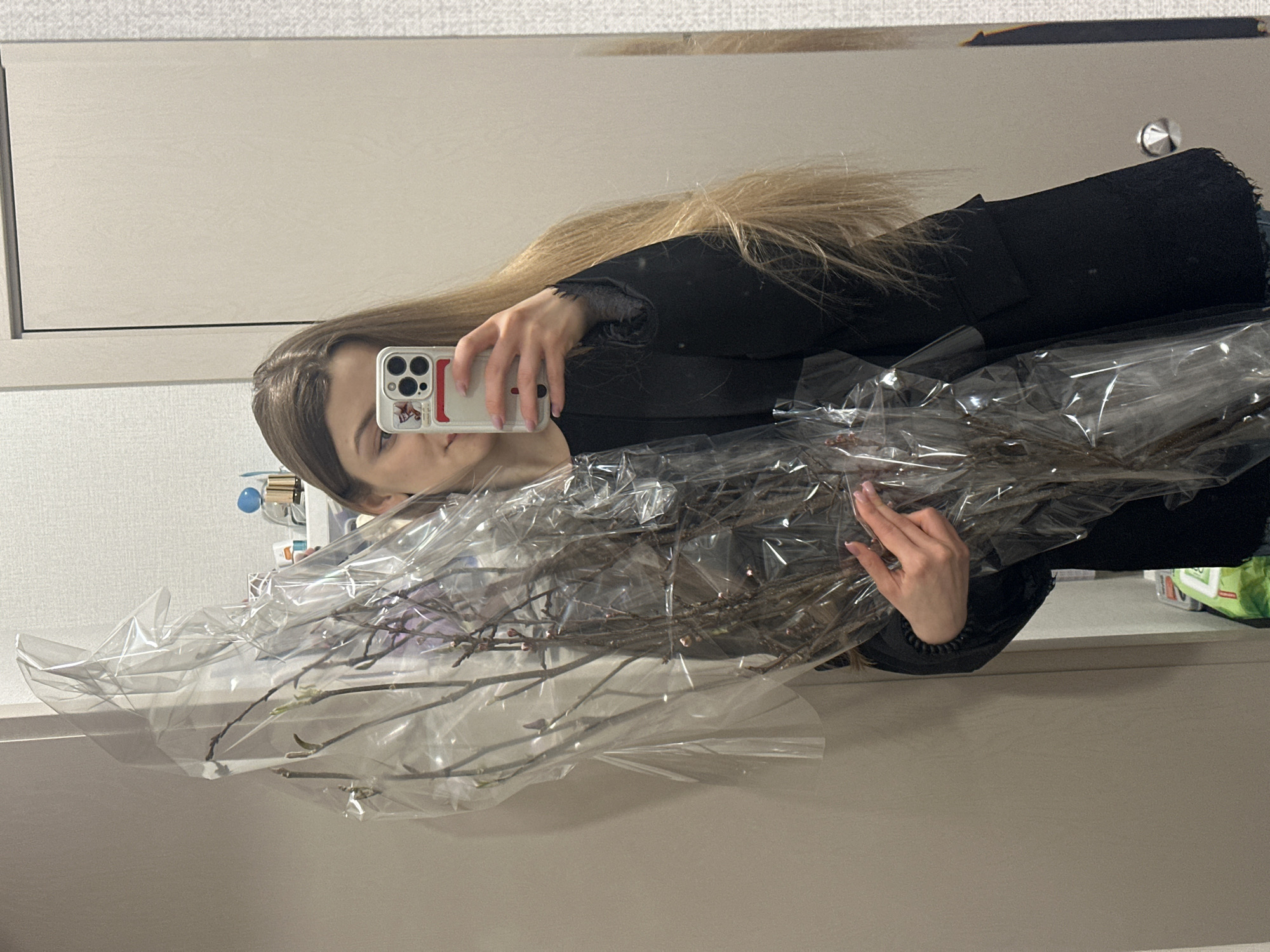 An indoor mirror selfie depicts a female individual holding a large bundle of dormant tree branches, wrapped in clear plastic film, within what appears to be a residential setting in Kazan, Russia. The subject's face is partially obscured by a white-cased smartphone, which she is using to capture the image. She has long, light-colored hair and is wearing a dark long-sleeved garment. Her right hand holds the smartphone, with visible light pink nail polish, while her left hand supports the lower portion of the wrapped branches. The background, visible in the reflection, includes a light-textured wall, a plain light-colored door or wardrobe panel featuring a chrome, conical doorknob, and a partially visible shelf with various small containers, along with a green bag near the floor.