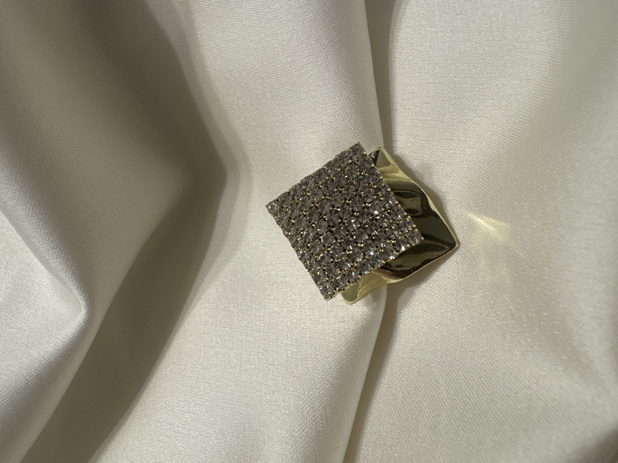 A close-up, eye-level shot shows a gold-colored ring adorned with small, clear stones. The ring is positioned on a draped surface of ivory-colored satin fabric, which exhibits subtle folds and shadows. The fabric's texture appears smooth and lustrous, reflecting soft light. The background is entirely composed of this draped fabric, with the focus sharply on the ring. No people or other objects are visible in the image. The image was taken indoors, likely in a studio setting, given the controlled lighting and the arrangement of the fabric. There is no information in the image to suggest a specific location within Bishkek City, Kyrgyzstan.