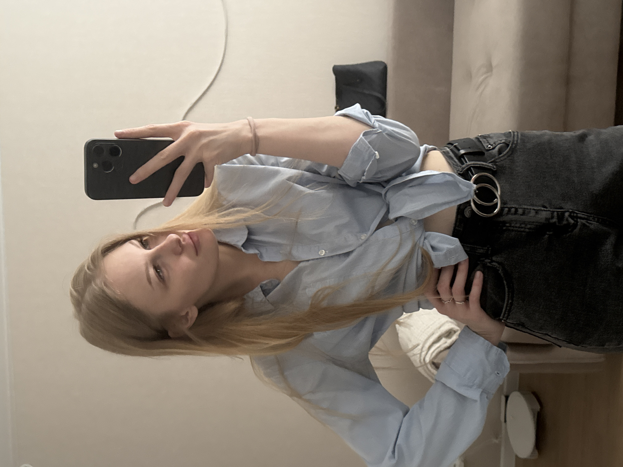 A young Caucasian woman with blonde hair, wearing a light blue button-up shirt tied at the waist and dark jeans with a double-ring belt, takes a mirror selfie in what appears to be a residential setting, possibly an apartment in Perm, Russia. She is standing, with her left hand on her hip and holding a dark-colored smartphone in her right hand. Her gaze is directed towards her reflection. In the background, a neutral-colored sofa and a dark bag are visible. The lighting suggests an indoor environment.