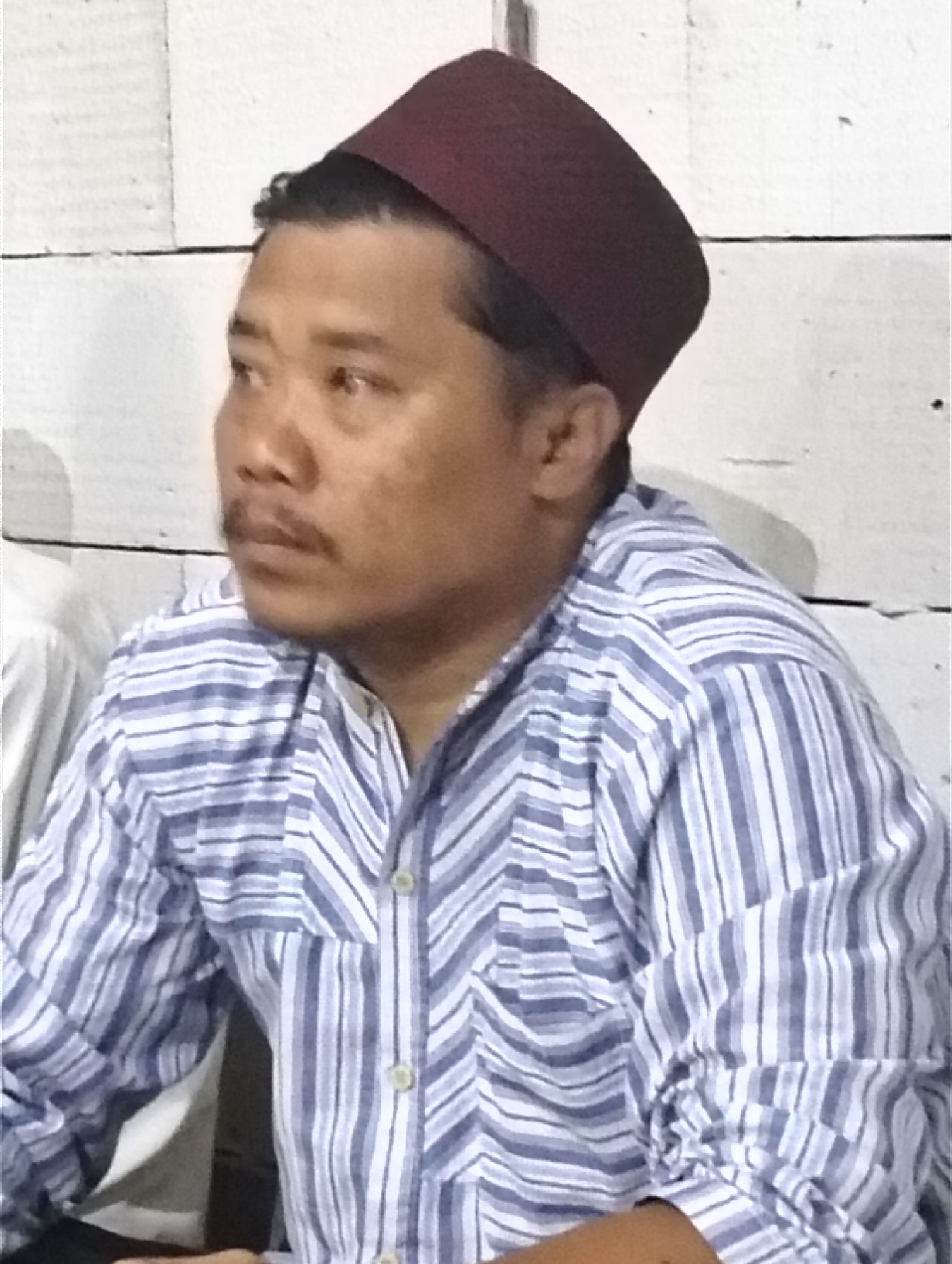 A man wearing a maroon kufi and a blue and white striped button-down shirt is shown from the chest up, looking to his left. The background is a white wall with a horizontal line across it. The man has dark hair, brown eyes, and a mustache. His shirt has two visible buttons. The image appears to be taken indoors.