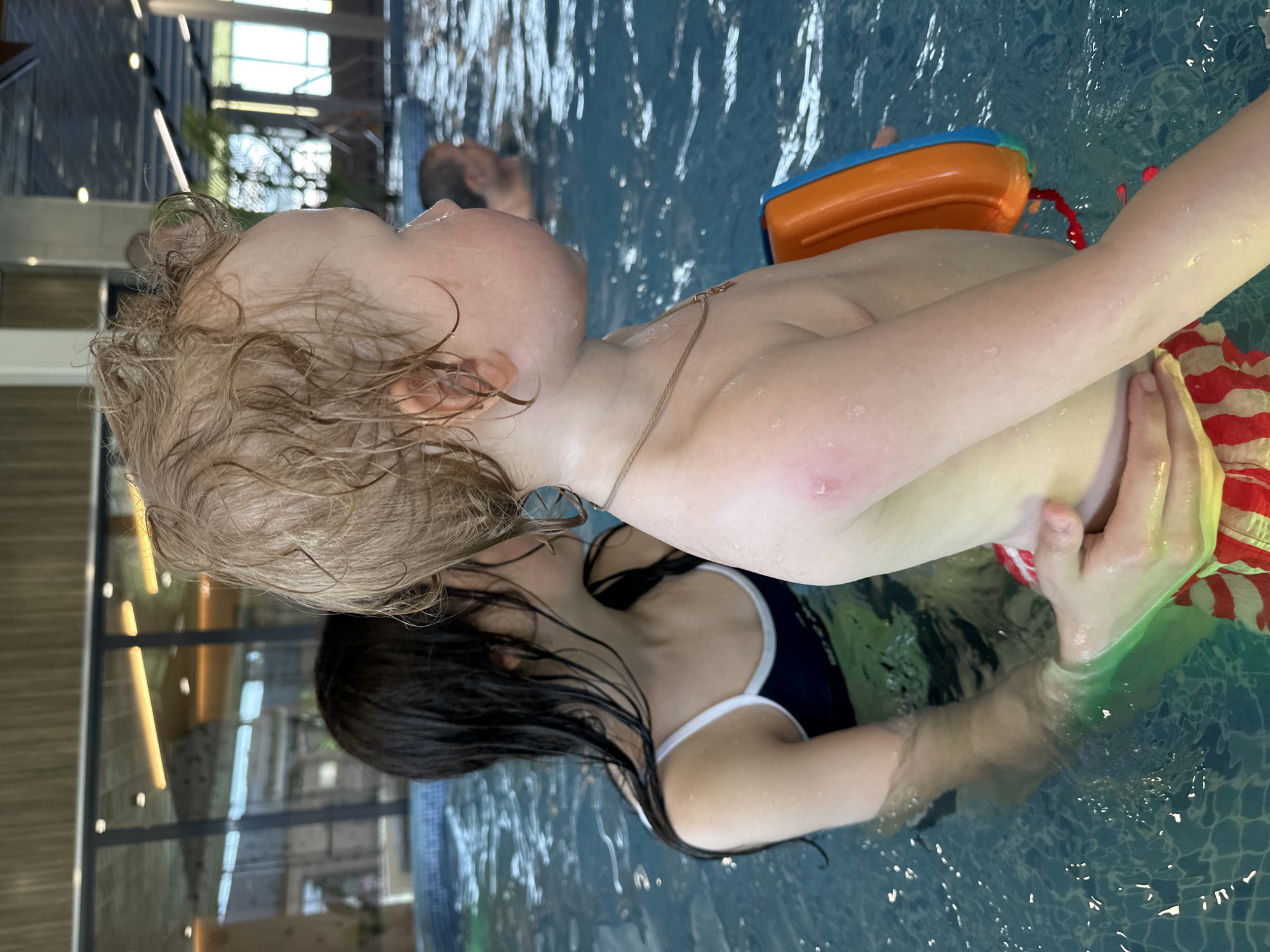 A woman is holding a child in a swimming pool. The child has blonde, wet hair, and is wearing a necklace and red and white striped shorts. They are also holding an orange and blue toy. The woman has dark, wet hair and is wearing a dark blue swimsuit with white trim. The pool water is blue and there are other people in the background. The location appears to be indoors, with large windows and supporting columns. The scene may be in Votkinsk, Russia.