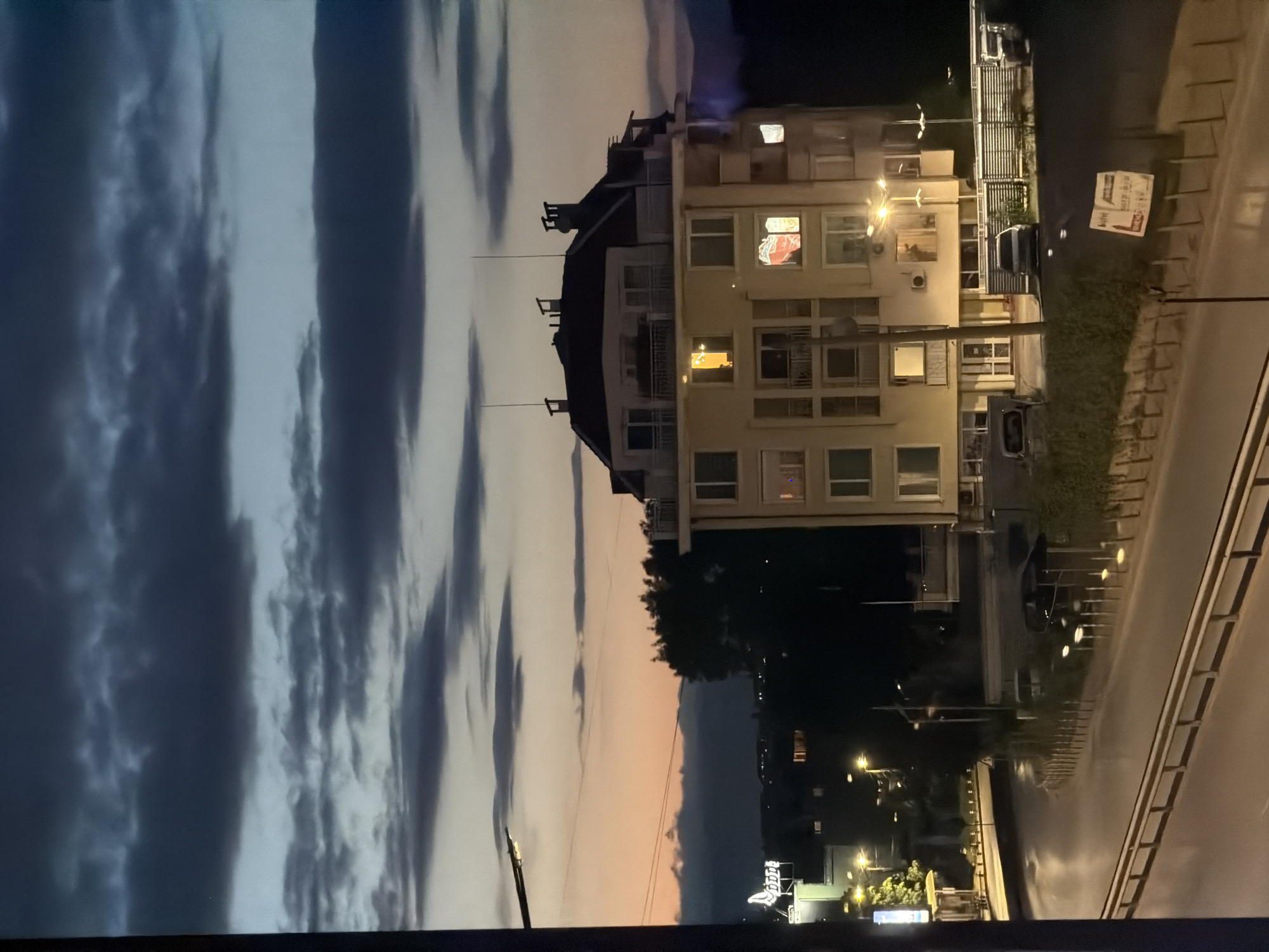 The image shows a residential building in Sofia, Bulgaria at dusk. The sky is a mix of dark blue clouds and a peachy sunset. The building is multi-story and lit with interior lights. A road runs alongside the building, with streetlights illuminating the scene. Other buildings and lights can be seen in the distance.