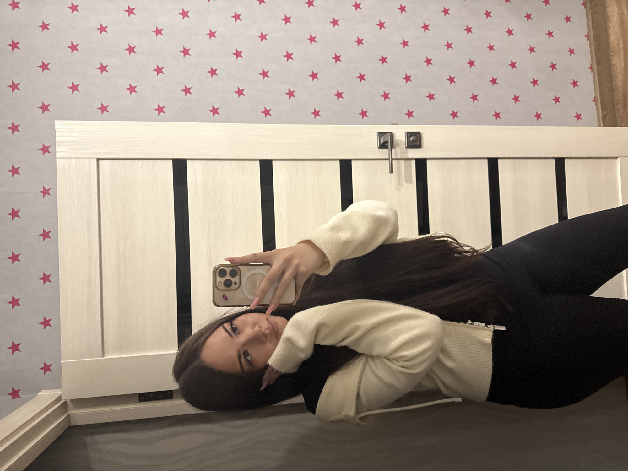The image displays a mirror selfie of a young woman in an indoor setting, likely a bedroom, in Naberezhnye Chelny, Russia. The photograph is rotated 90 degrees clockwise. The woman has long, dark hair, dark eyebrows, and is wearing a cream or off-white zip-up sweater and dark leggings. She holds a light-colored smartphone with a multi-lens camera array and a small sticker on its case, reflecting her face and upper body as she poses. Her expression is calm and neutral.
The background features light gray or off-white wallpaper decorated with numerous uniformly spaced pink five-pointed stars. Below the wallpaper, a light-colored wooden structure, possibly a headboard or a piece of furniture, is visible, characterized by vertical dark slats and two dark metallic handles or fixtures on its top edge. A fragment of a light brown curtain is visible on the far right. The indoor lighting appears bright and even, without indicating a specific time of day. There is no visible text in the image.