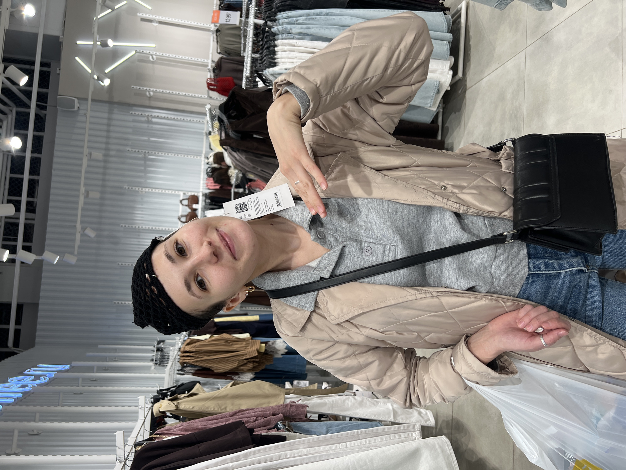 The image captures a young woman inside a clothing store in Perm, Russia, rotated 90 degrees counter-clockwise. She is the central subject, looking directly at the camera with a subtle smile. She wears a black knitted headband, a light grey long-sleeved shirt, and a beige quilted jacket. A black shoulder bag with a textured flap crosses her body. Her left hand holds a white plastic shopping bag, while her right hand gestures towards her chest, displaying a clothing tag that reads "H&M" and includes a barcode and the currency "EUR." A silver-colored ring is visible on her left ring finger.
The background shows typical elements of a retail clothing environment. Racks of folded and hanging clothes, including denim and various colored garments, line the walls. Bright, modern fluorescent lighting fixtures are mounted on the ceiling and walls. White shelving units and some dark-colored handbags or accessories are also visible. The floor is light-colored and tiled. On a red tag attached to an item on a distant rack, the number "1299" is partially legible. The overall lighting is bright and even, characteristic of indoor commercial spaces.
