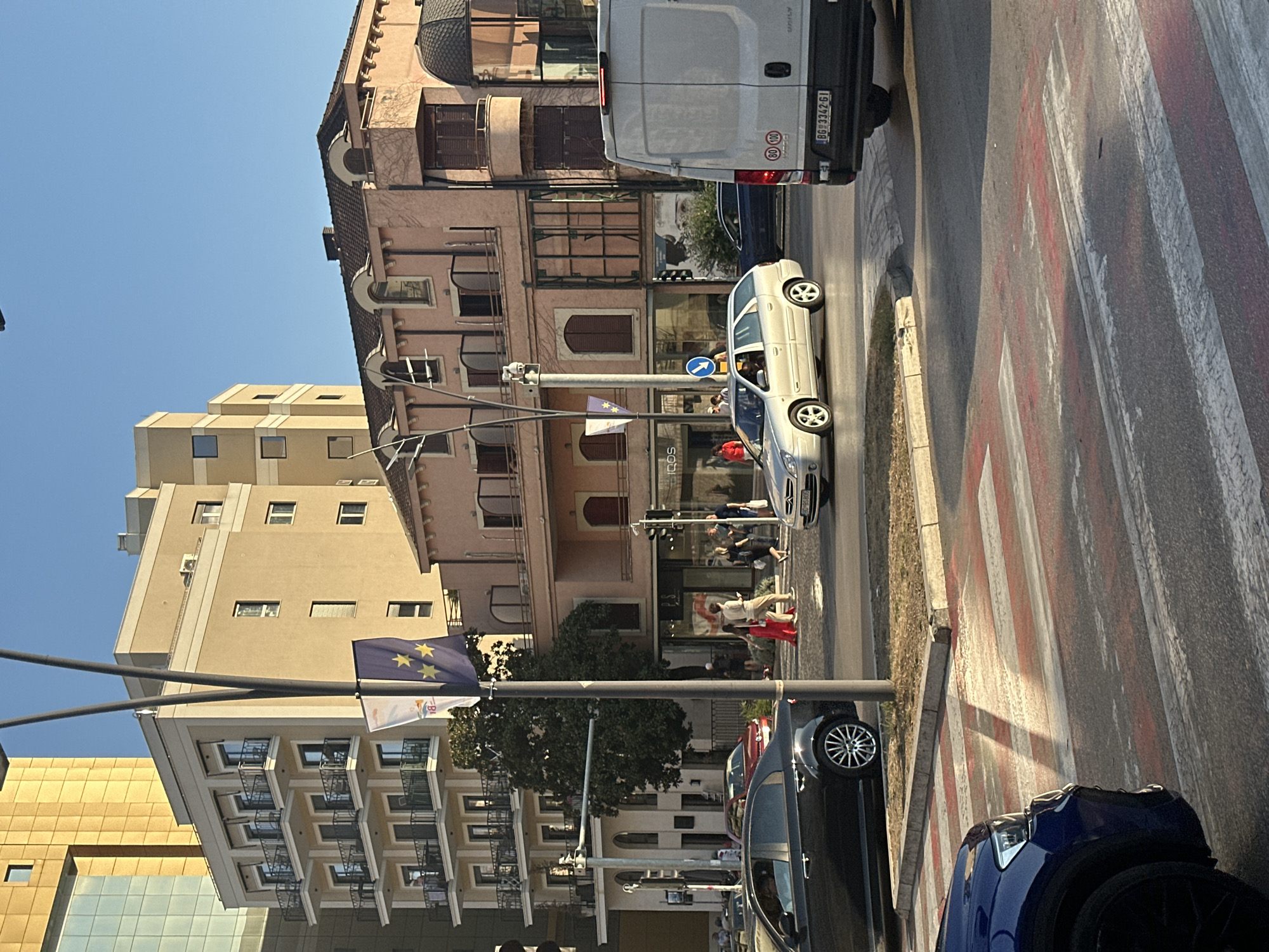 The image captures a street scene in Budva, Montenegro, bathed in the warm light of the late afternoon sun. In the foreground, a blue SUV is partially visible, its dark paint reflecting the light. Across the street, a pedestrian crossing with red and white stripes marks a designated area for walkers.
Several cars are in motion and parked. A black sedan is positioned on the left side of the road, its front wheel turned. Further down the street, a silver station wagon is in the process of turning, with its driver visible behind the wheel. A white van with "DAILY" branding on its rear door is parked on the right side of the road, its license plate reading "BG 3342-GI". A red car is parked further back in the scene.
Buildings of various architectural styles line the street. A modern, golden-hued building with glass facades stands on the far left. Adjacent to it is a lighter-colored building with multiple balconies and numerous windows. To its right is a pink building with arched windows and shutters, and a darker tiled roof. Another building to the far right has a dark domed section.
Several people are visible on the sidewalks and crossing the street. Two individuals in the center are walking across the pedestrian crossing, one wearing a red dress. Other pedestrians are seen on the sidewalks, some near storefronts. Traffic lights and streetlights are present, indicating an urban intersection. Two flags, one white and one dark blue with yellow stars, hang from a tall pole in the middle ground. The sky is clear and blue, suggesting fair weather.