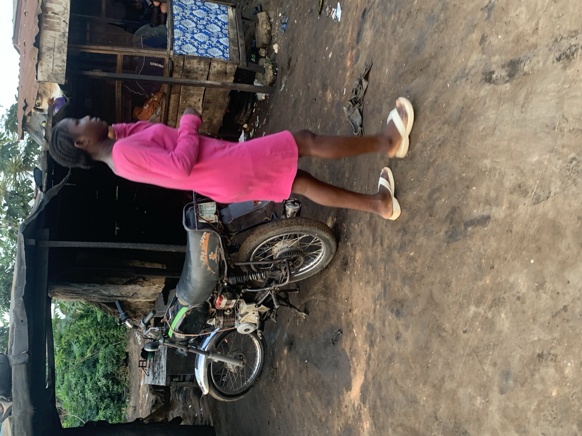 A young Black female wearing a bright pink, long-sleeved dress and white slip-on sandals walks past a parked motorcycle in an outdoor setting, likely in Shagamu, Nigeria. The motorcycle, a dark color with a green stripe on the fuel tank, is positioned to the left of the frame. The ground is a dusty, unpaved surface. Behind the motorcycle is a rustic structure, possibly a stall or shop, with a corrugated metal roof and wooden walls. Lush green foliage is visible in the background. The lighting suggests it is daytime, possibly late afternoon.