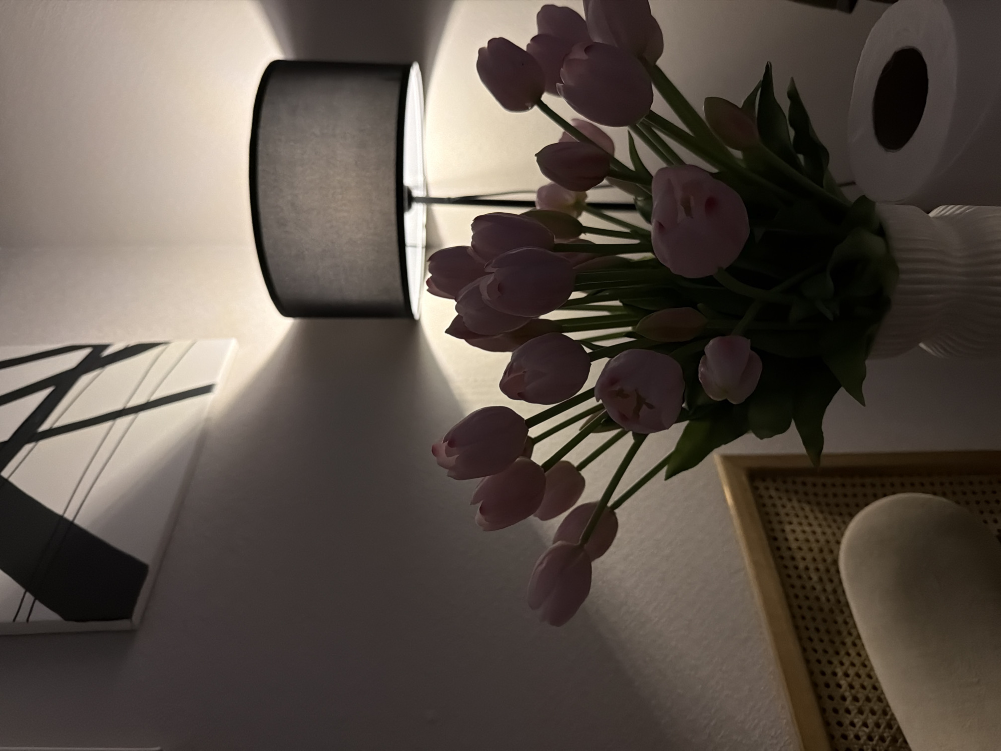 In Moscow, Russia, a close-up shot captures a dimly lit room, possibly a bedroom or living area. A white vase filled with pink tulips sits on a surface, illuminated by the warm glow of a black lampshade. The lamp, positioned behind the flowers, casts a soft light, creating shadows on the wall. To the left of the tulips, a piece of abstract art with black lines on a white background is visible. The foreground shows a portion of a cane-backed chair and what appears to be a roll of toilet paper to the right of the vase. The overall ambiance is serene and intimate.