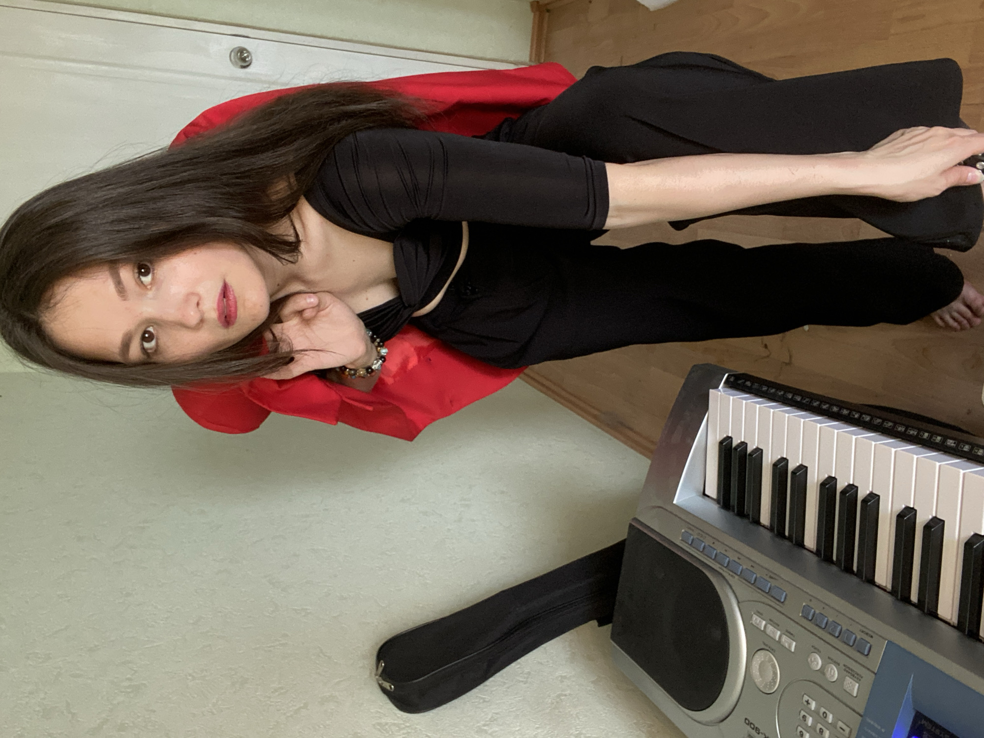 In this media file, a young woman with long, dark hair lies on a red fabric, possibly a cape or shawl, draped over a chair. She wears a black, long-sleeved top and black pants. Her head is tilted, and she gazes upwards with a thoughtful or pensive expression. Her left hand rests near her face, adorned with a beaded bracelet, while her right arm is extended along her leg.
Beside her, a keyboard with white and black keys is partially visible, suggesting a musical setting. A black case for the keyboard lies on the floor. The background consists of a light-colored wall and wooden flooring. The lighting appears to be indoor, possibly from natural light as there are no harsh artificial shadows.
The image is oriented sideways. No specific time of day or weather conditions are discernible. The presence of the keyboard hints at a music-related activity or hobby. The woman's pose and expression could convey a sense of relaxation, creativity, or contemplation. No visible text is present in the image. The location context provided is Chaikovsky, Russia.