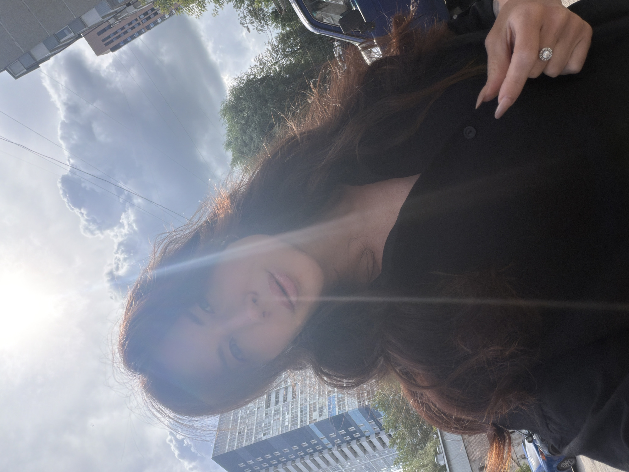 A woman with long, auburn hair and a black shirt takes a selfie outdoors. She is wearing a ring on her right hand, which is visible near the bottom right of the frame. In the background, there are several tall buildings, trees, and a blue vehicle. The sky is partially cloudy with the sun shining brightly, creating lens flare. The architecture suggests an urban environment, possibly a residential or business district. The overall impression is that of a casual, sunny day in a city.
The image was taken in Perm, Russia. The tall building in the background with a blue facade and white balconies is characteristic of residential complexes built in Perm during the late Soviet or post-Soviet era. The presence of power lines and the style of the buildings are also consistent with urban areas in Russia. The blue vehicle could be a common model found in Russia, such as a Lada or a foreign car popular in the region.