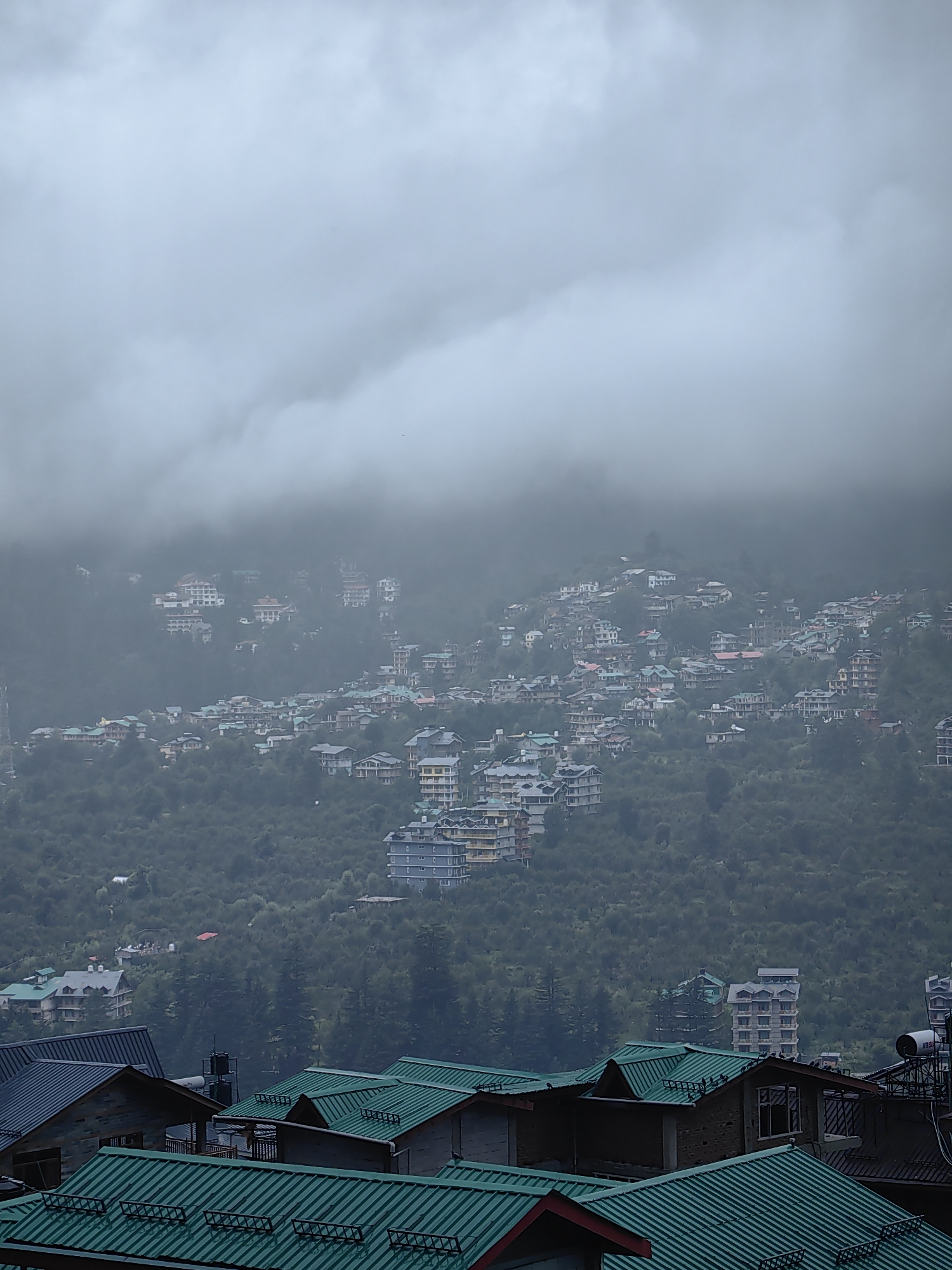The image depicts a dense hillside settlement in Vashisht, India, under an overcast, cloudy sky. The terrain is steep, with numerous buildings constructed at varying levels, nestled amongst lush green trees. The architecture is varied, featuring multi-story structures with balconies and flat roofs, as well as smaller houses with peaked, green-tiled roofs in the foreground. The buildings are tightly packed together, suggesting a well-developed urban area on the mountainside. The cloudy weather creates a muted, diffused light across the scene, with some clouds obscuring the upper portions of the hills and the sky. The foreground is dominated by several buildings with prominent green, corrugated metal roofs, showing architectural details like window frames and supporting structures. Some of these buildings appear to be residential or commercial. The overall impression is of a bustling town built into the natural landscape, facing a subdued, possibly rainy, day. There are no visible people or animals in the image.