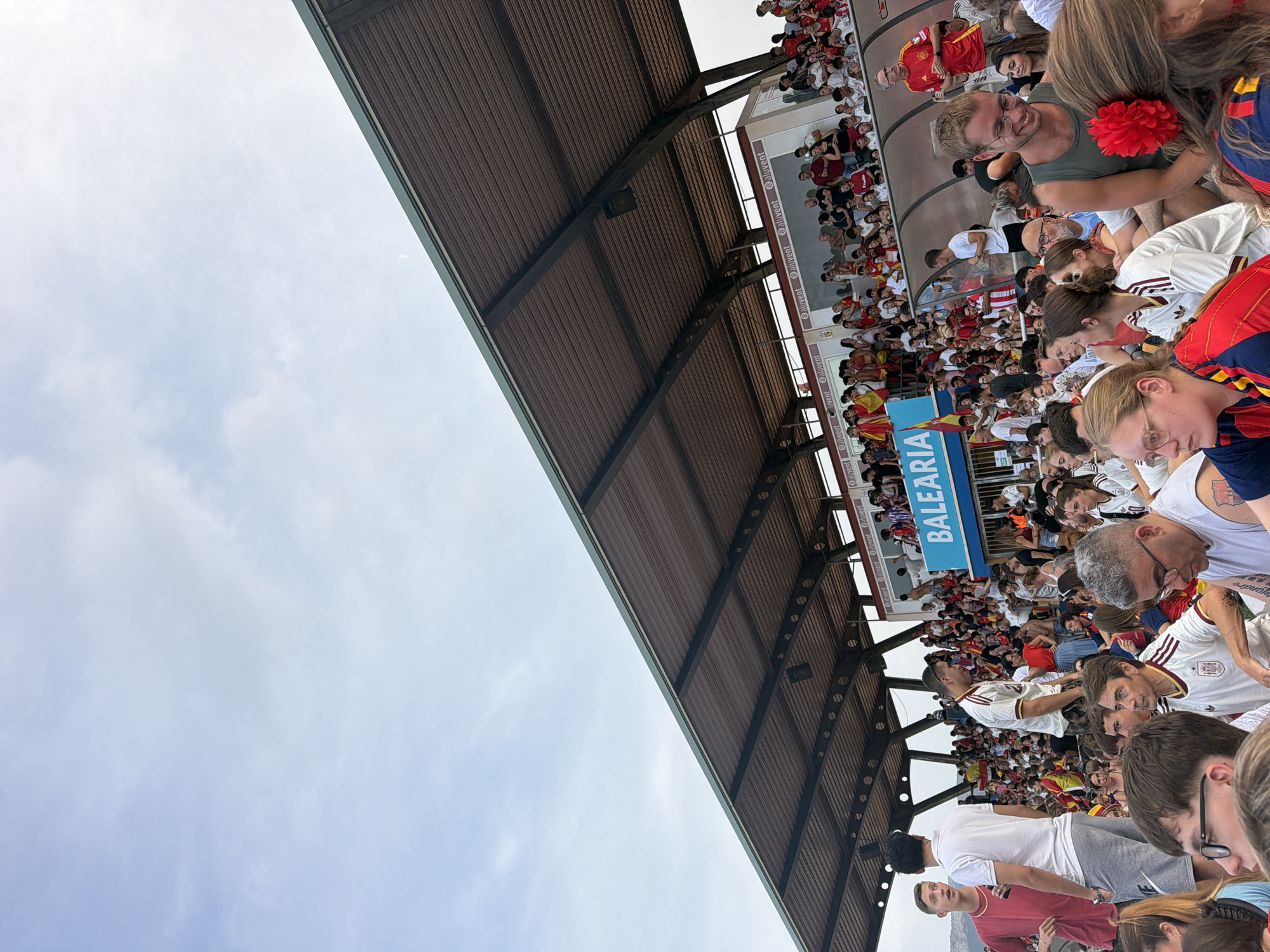 The image depicts a crowded outdoor stadium seating area, likely during a sporting event given the presence of fans wearing team colors and holding flags. In the background, a large banner reads "BALEARIA" in blue and white lettering, indicating a sponsor or location. The stadium appears to be a modern facility with a covered roof structure supported by metal beams. Spectators are seated and standing, some are interacting with each other, and others are looking towards an unseen event area. The overall atmosphere suggests a lively and engaging event. The presence of the "BALEARIA" banner, a ferry company operating between the Balearic Islands and the Spanish mainland, strongly suggests the location is the Balearia Stadium in Dénia, Spain.