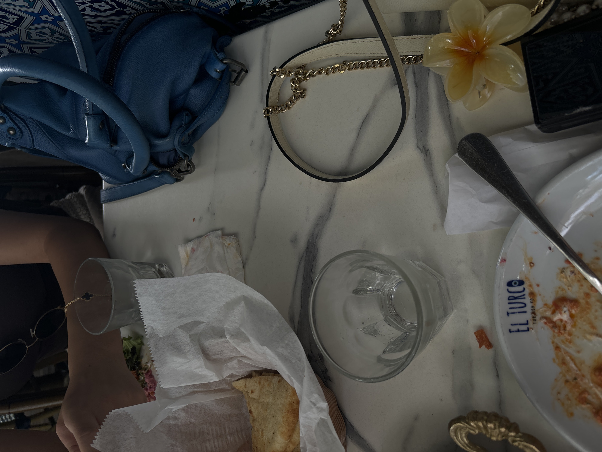 The image shows a marble table at a restaurant, possibly El Turco Turkish Food (as indicated by a plate) in Miami, Florida. There is a blue leather purse, a white purse with a chain strap, a decorative flower, a half-eaten plate of food with a spoon, an empty glass, a sandwich wrapped in paper, sunglasses, and a person's arm with a bracelet. The overall scene suggests a casual dining experience.