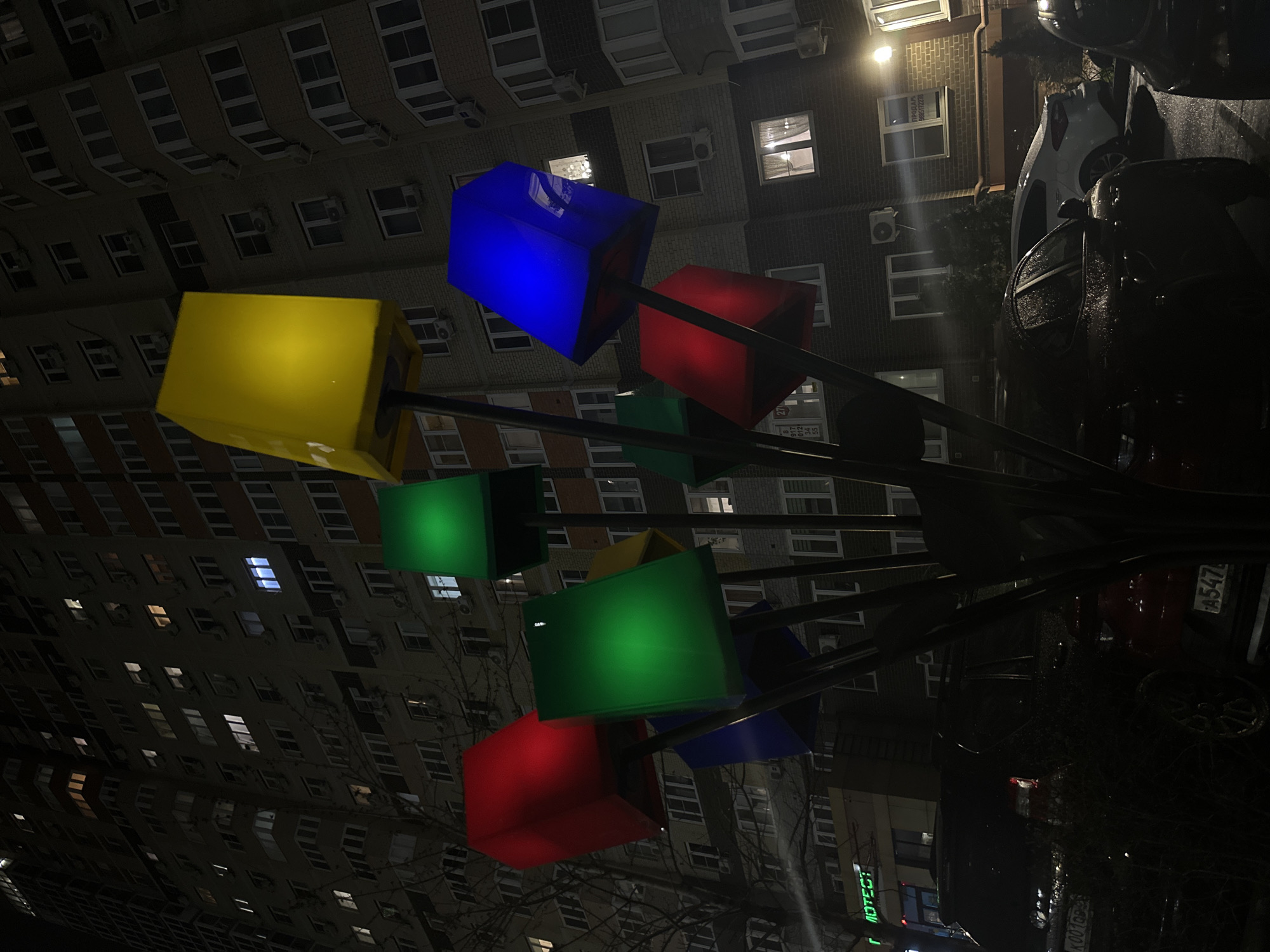 The image captures a nighttime scene in Rostov-on-Don, Russia. A colorful art installation featuring illuminated cubic shapes of yellow, blue, red, and green stands prominently in the foreground. The structures are mounted on metal poles that branch out like stems. In the background, a multi-story residential building looms with rows of windows, some lit. Parked vehicles are visible on the right side, including a dark-colored car with a visible license plate "A547A". The overall ambiance suggests a rainy or wet evening, given the reflections on the car surfaces and wet road. The scene appears to be an urban landscape where the artwork blends with the everyday environment of buildings and transportation.