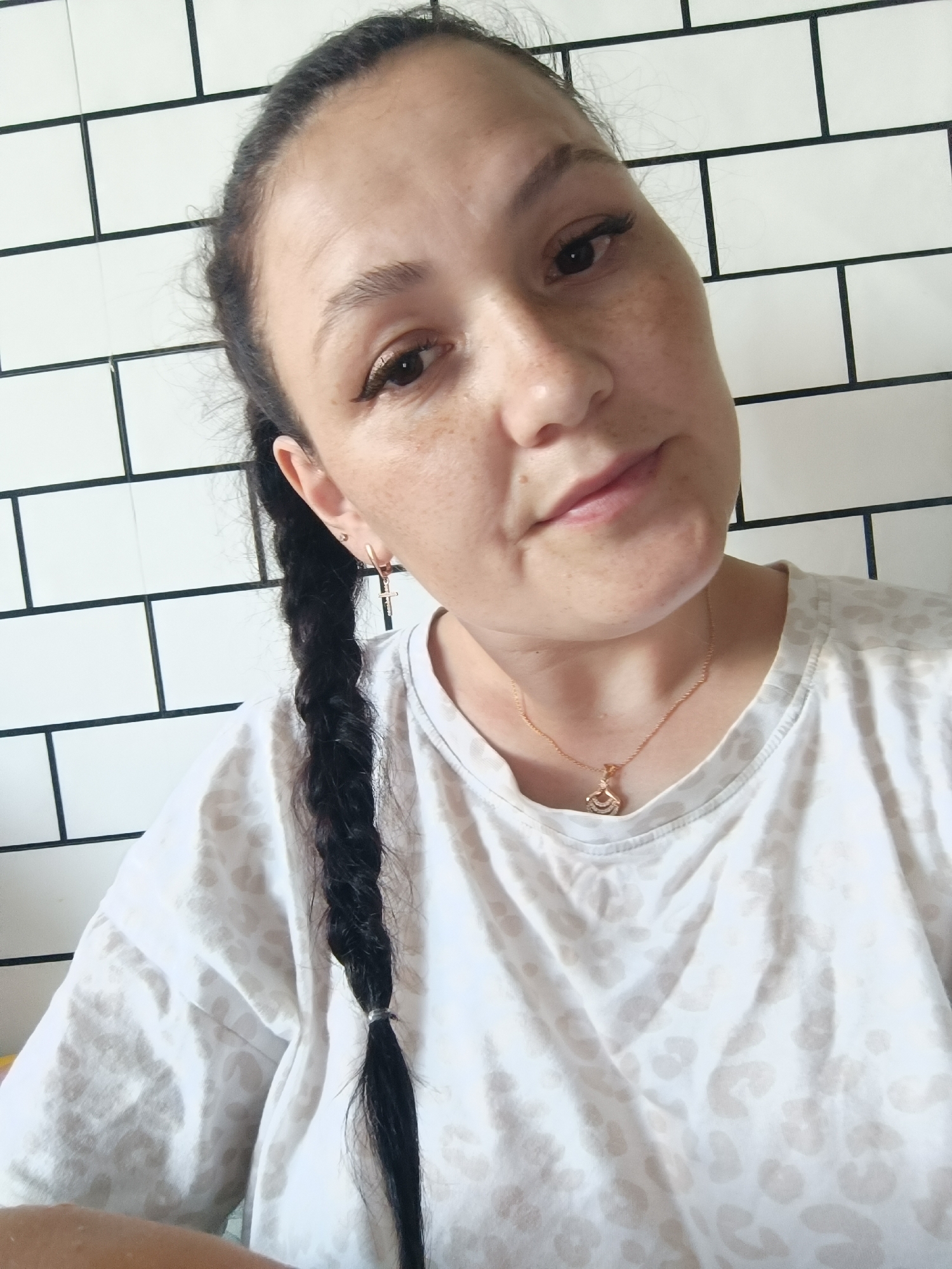 A selfie is taken by a woman with dark hair in a braid, in front of a white tiled wall. She is wearing a white t-shirt with a leopard print pattern and a gold necklace with a pendant. The woman has freckles on her face. The setting appears to be indoors. There is no indication of the city of Chaikovsky, Russia.