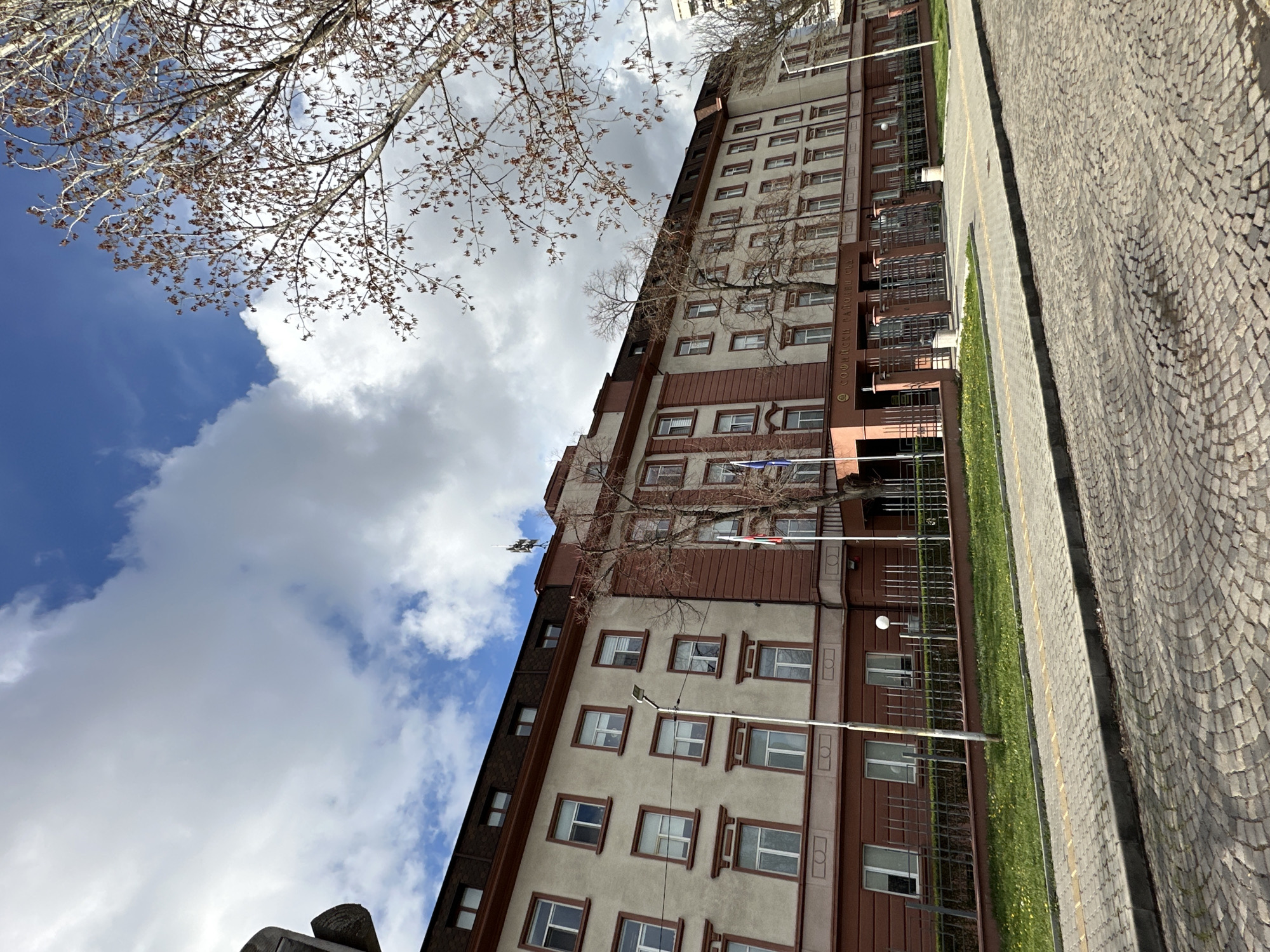 A low-angle shot shows a tall, multi-story building against a cloudy blue sky. The building is light beige with brown accents around the windows and along the edges. It appears to be in Sofia, Bulgaria. Trees with bare branches are visible in the foreground. To the right of the building is a sidewalk or street paved with cobblestones. A strip of green grass separates the building from the paved area. Several flagpoles with flags are erected near the building's facade. The sky features a mix of blue patches and large, puffy white clouds.