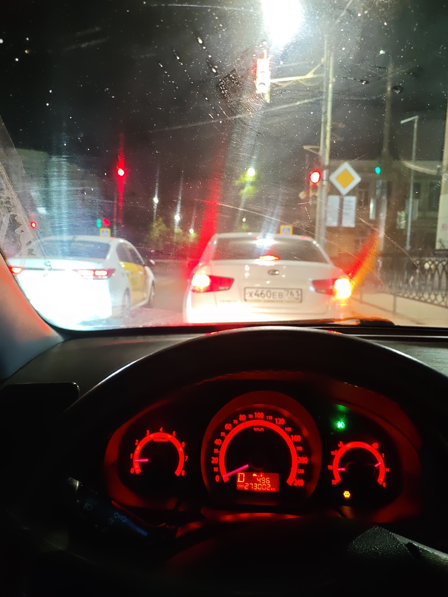 This media file depicts a view from inside a car at night, likely in Rostov-on-Don, Russia, due to the visible license plate format. The driver is stopped at a traffic light, indicated by the red lights illuminated ahead. The windshield is wet, suggesting recent rain or ongoing precipitation.
The foreground shows the dashboard of the car, illuminated by the red glow of the instrument cluster. The speedometer shows a speed of 0 km/h, and the odometer reads 273002 km. A green indicator light is illuminated on the dashboard.
Outside the car, several vehicles are visible. To the left, a white car, possibly a taxi due to the yellow stripe, is also stopped. Ahead, a white sedan with a license plate reading "X460EB 761" is also waiting. Traffic lights and road signs, including a yellow diamond-shaped sign, are visible in the distance. The scene is illuminated by streetlights and the headlights of other cars, creating reflections on the wet windshield. The overall atmosphere is one of a typical nighttime urban commute during inclement weather.