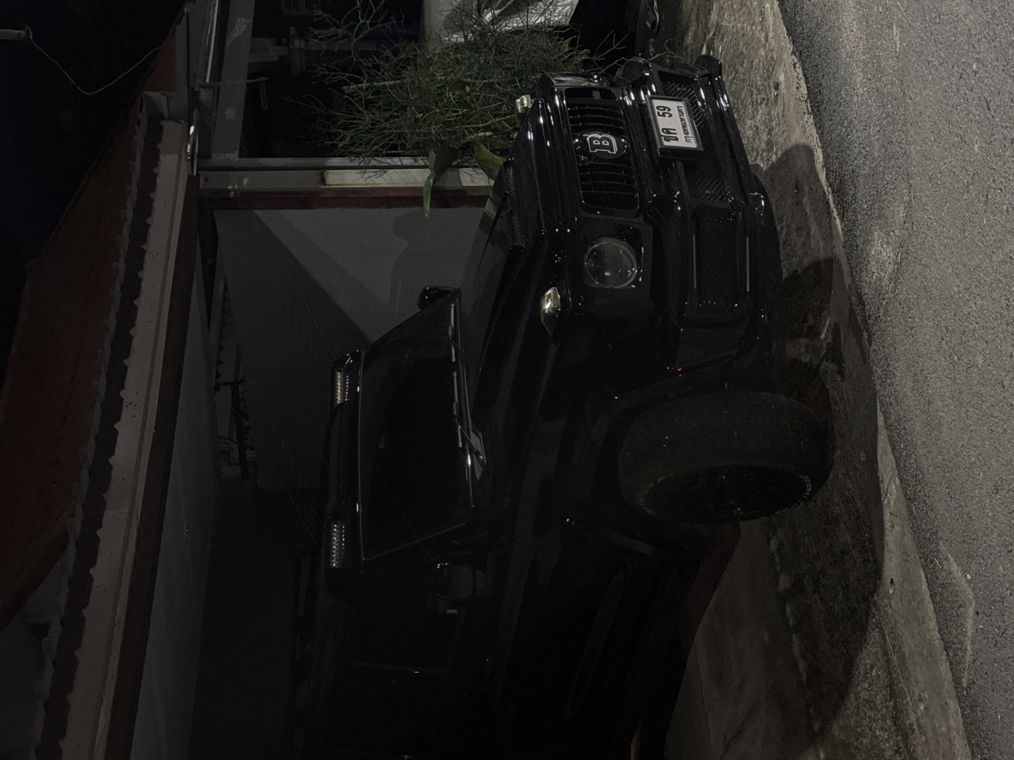 A black Mercedes-Benz G-Class SUV is parked in an open garage at night. The vehicle is glossy black and features aftermarket LED light bars on the roof, a custom front grille with a "B" emblem, and a modified front bumper. The license plate, which appears to be from Thailand, reads "ขด 59" and "กรุงเทพมหานคร". The car is positioned at an angle, with its front end facing slightly towards the viewer and to the right. The garage has a corrugated metal roof and concrete walls. To the right of the car, a dense bush with thin, branch-like foliage is visible. The ground in front of the garage is asphalt, with some puddles or wet patches. The lighting is dim, suggesting it is nighttime, with artificial light illuminating the scene. No people are present in the image.