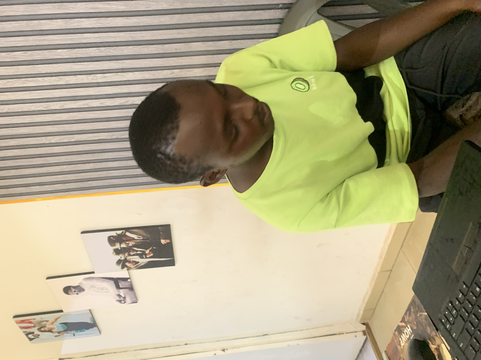 An image depicts a person with dark skin, wearing a lime green long-sleeved shirt with a "Kiwi" logo, sitting indoors. The person appears to be looking down, possibly at a laptop keyboard visible in the foreground. The indoor environment includes a wall with three framed pictures, and horizontal wooden slats overhead. There is insufficient information to definitively place the scene in Jos, Nigeria.
