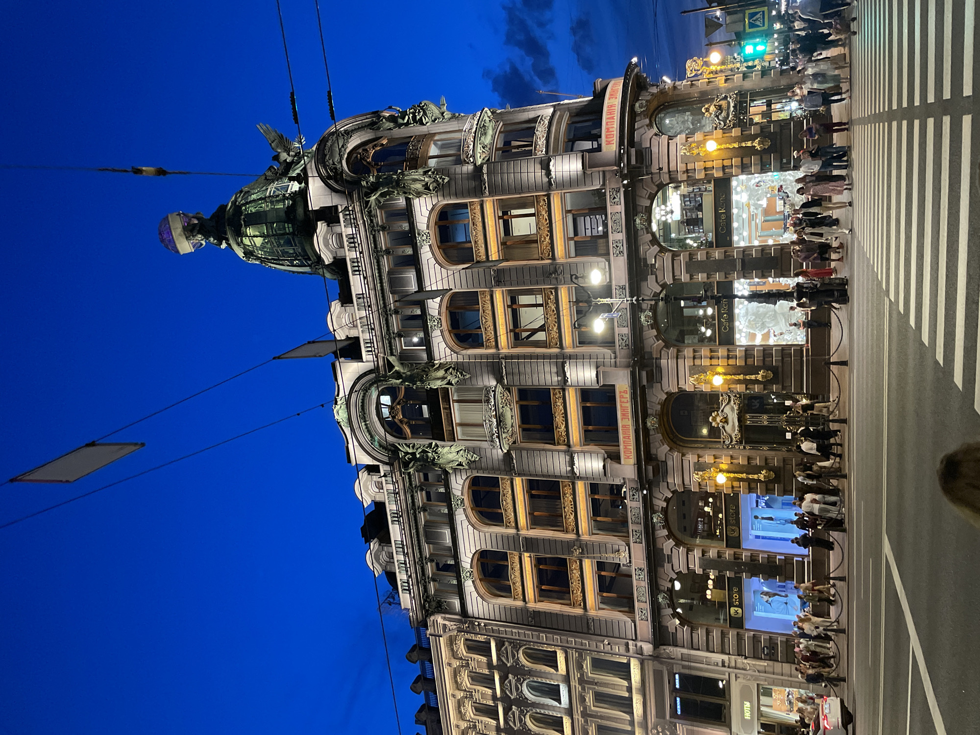 This image captures a vibrant evening scene at the Singer House (Dom Knigi - House of Books) in Saint Petersburg, Russia, specifically at the intersection of Nevsky Prospekt and the Griboyedov Canal Embankment.
The prominent structure is the multi-story Singer House, illuminated against a deep blue twilight sky. The building, designed in a Neo-Baroque style, features a distinctive green dome topped with a glass globe, which emits a purple light. An eagle statue is visible on the dome's facade. The building's exterior is light-colored, adorned with intricate architectural details, including ornate window frames, balconies, and decorative elements, many of which are highlighted by external spotlights. The inscription "КОМПАНИЯ ЗИНГЕРЪ" (Company Singer) is visible on the lower part of the facade. Storefronts on the ground level include a "vk store" with brightly lit digital displays and mannequins, and "Cafe René" with reflective windows and white interior decorations. Another establishment, "НОТЫ" (Notes), is partially visible to the far left.
Overhead utility wires crisscross the sky, with a rectangular banner hanging from them in the upper left quadrant. Streetlights with warm yellow illumination are positioned along the sidewalk.
At street level, a significant number of pedestrians are present. Dozens of individuals and small groups, dressed in casual clothing, are seen walking along the wide sidewalk and actively crossing a clearly marked zebra pedestrian crossing in the foreground. They move in various directions, indicating a busy urban environment. A white passenger vehicle is partially visible on the far left edge of the street. Traffic lights are visible on the right, with a green light indicating a pedestrian crossing signal. The road surface is asphalt, with distinct white markings for the crosswalk. The overall scene depicts a bustling city street during the blue hour, with architectural grandeur and active pedestrian life.