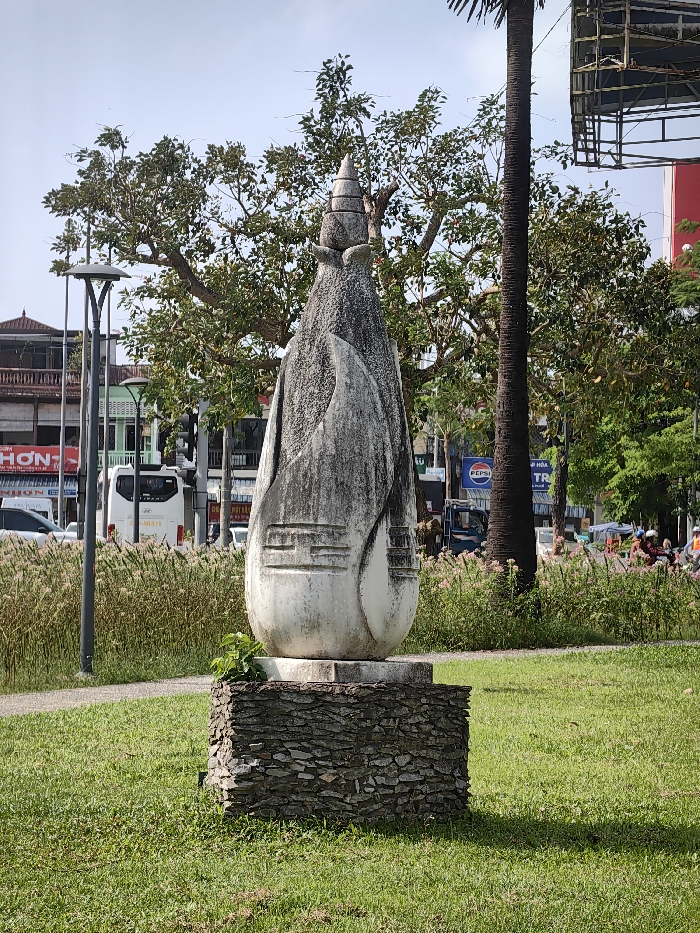 The image depicts an outdoor urban scene in Thuận Hóa Ward, Vietnam, under clear daylight conditions.
In the foreground, a tall, weathered, light-colored stone sculpture stands centrally on a rectangular base constructed from stacked, dark, irregular stones. The sculpture features an elongated, abstract, bulbous form, tapering to a multi-tiered, conical finial at its apex. Its surfaces exhibit significant grime and discoloration. Carved relief patterns, including horizontal lines and geometric shapes, are visible on its lower half. Small green plants are growing from the top edge of the stone base on the left side. The sculpture rests on a manicured green lawn, which casts a shadow to the right.
Behind the sculpture, a band of tall, wispy, light-brown grasses or reeds separates the lawn from a paved roadway. A dark-grey, multi-headed street lamp with a curving arm is positioned to the left, rising from these grasses.
The midground and background reveal a bustling street environment. Numerous mature trees with dense green foliage provide a natural backdrop, including a slender, dark-barked palm tree trunk on the right. Beyond the trees, multi-story urban buildings line the street, displaying typical Vietnamese architecture with balconies and storefronts. Visible signage includes "HON" and "NHON" on the left, and a "PEPSI" logo partially visible on the right. Traffic is present on the road, with a white passenger van (bearing "403.11") moving from left to right, and several motorbikes and scooters with riders further down the road to the right. A large metal scaffolding structure or billboard frame is partially visible above the trees on the far right. The sky overhead is clear and light blue.