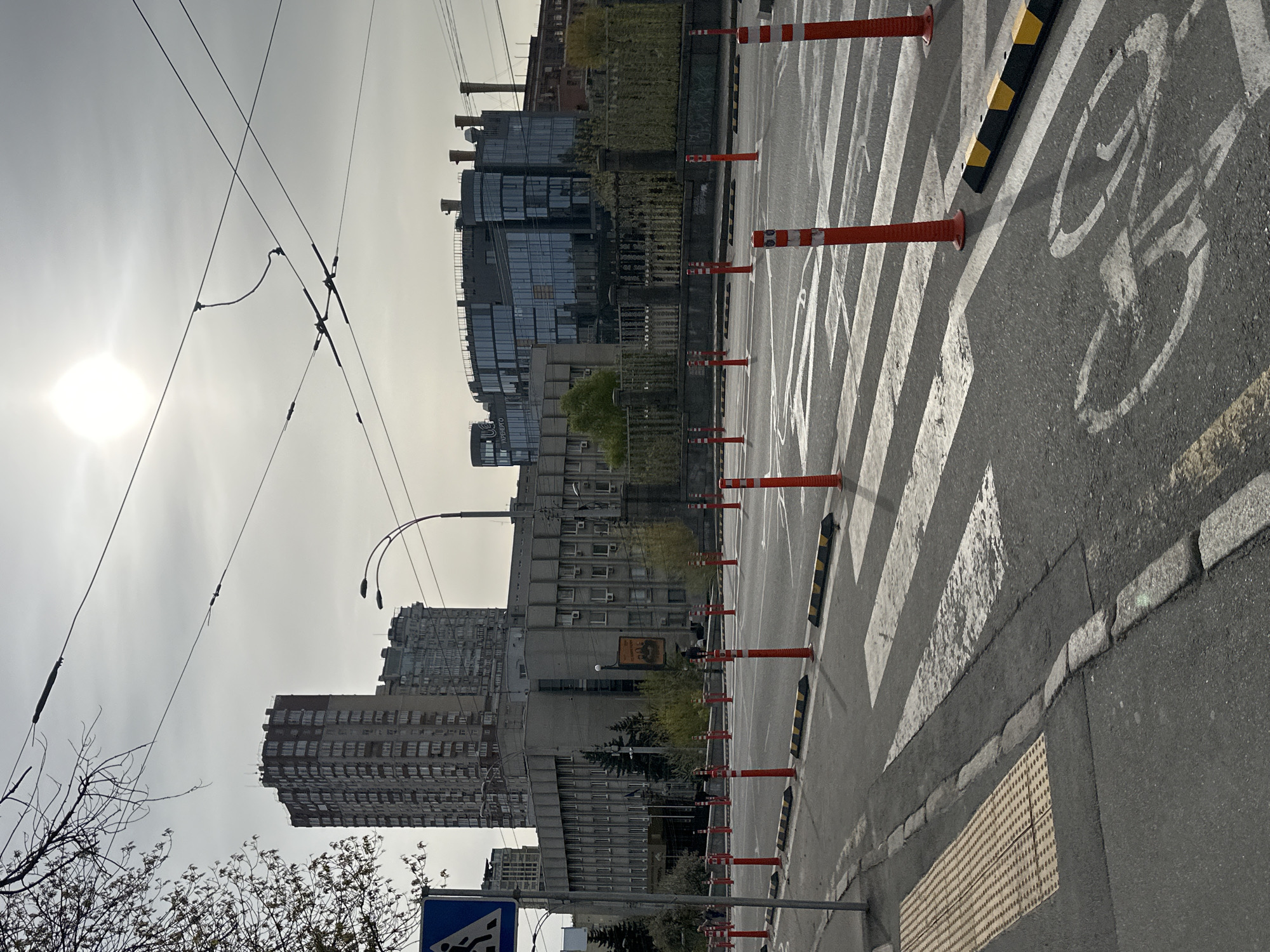 The image captures a street scene in Kyiv, Ukraine. The view is taken from the side of a road with a bike lane marked by a white bicycle symbol. The lane is lined with orange traffic bollards with white stripes. There's a pedestrian crossing with white stripes.
In the background, there are multiple buildings of varying heights and architectural styles. The nearest building is a high-rise apartment complex with numerous windows. A modern glass-faced building is also visible. Power lines crisscross the sky above the buildings.
The sky is overcast, with the sun partially obscured. Bare branches of a tree are visible in the bottom left corner, suggesting it may be late autumn or early spring. A blue and white road sign is visible.